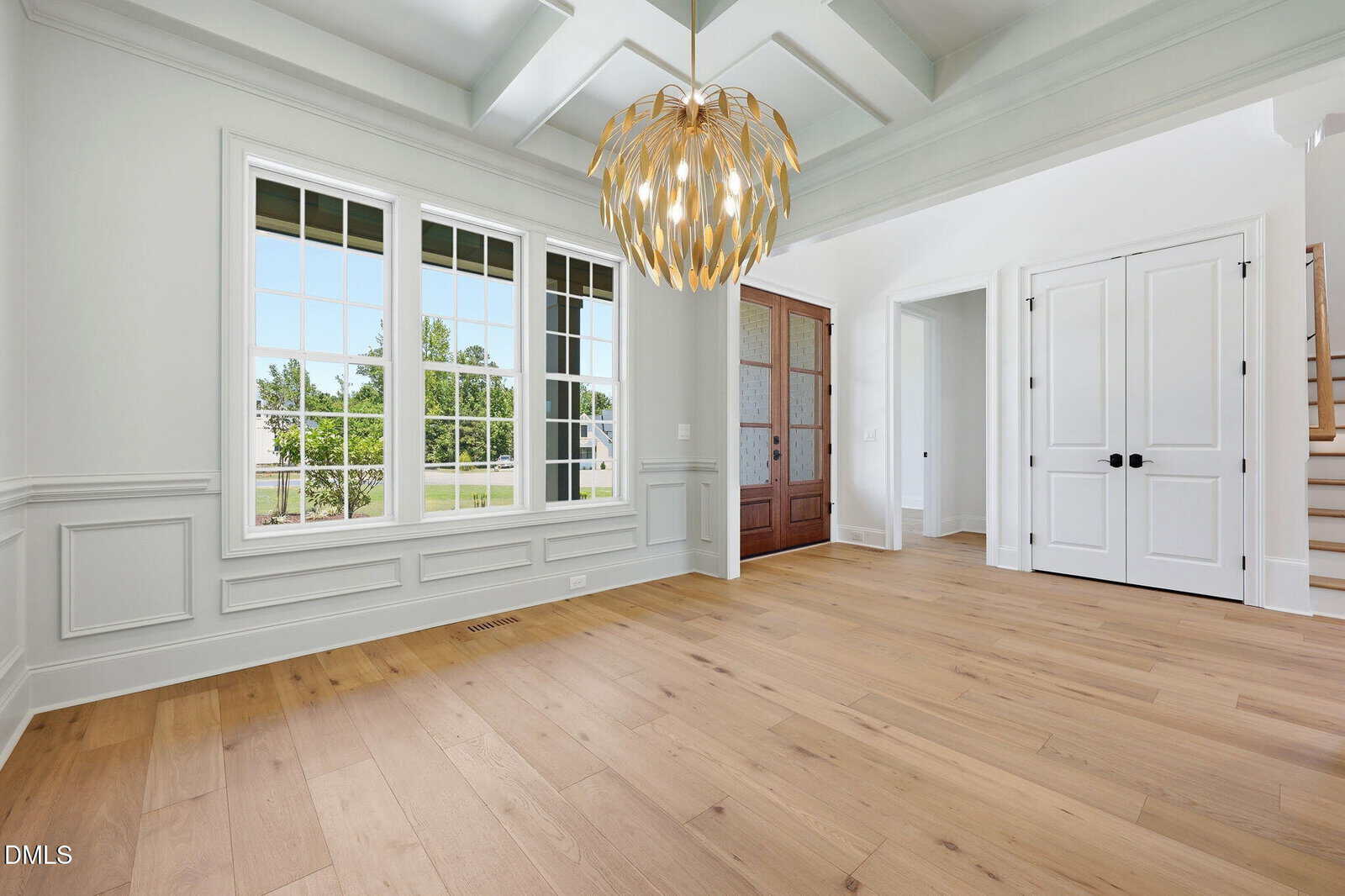 Bright, empty room with large windows, light wood floors, white paneled walls, and a modern chandelier. Wooden front door and staircase are visible, along with double white closet doors.