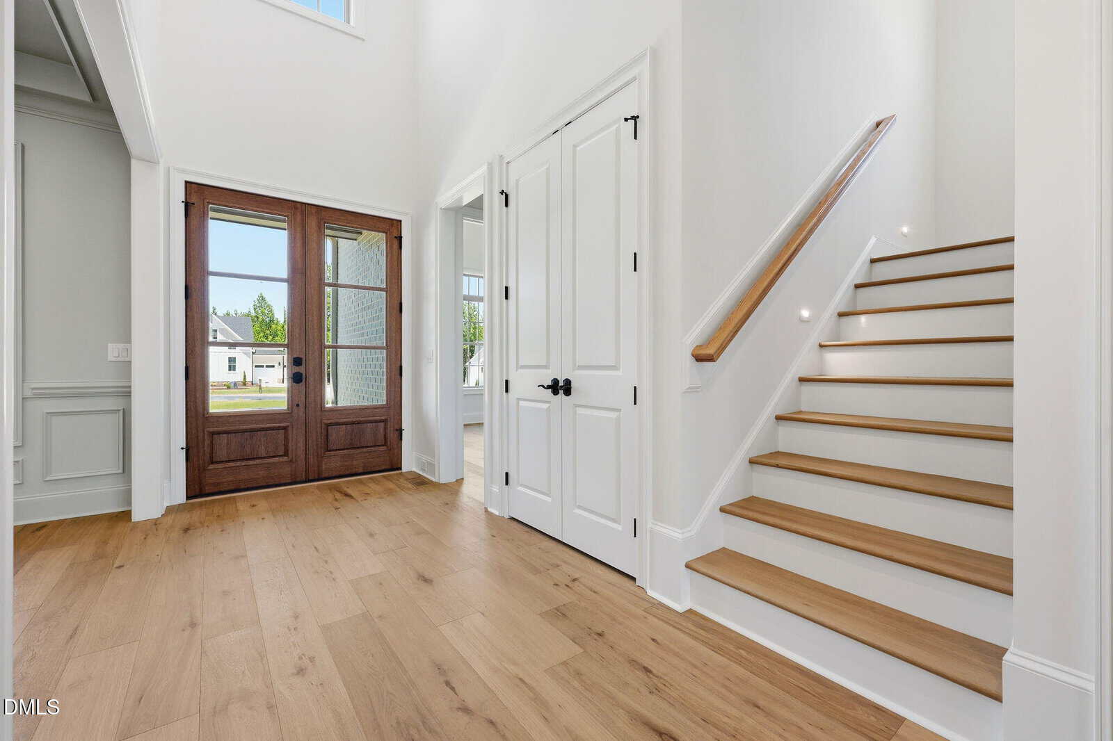 Bright entryway with light wood flooring, white walls, double wooden front doors with glass panels, a closet with white double doors, and a staircase with wooden steps and a simple handrail.