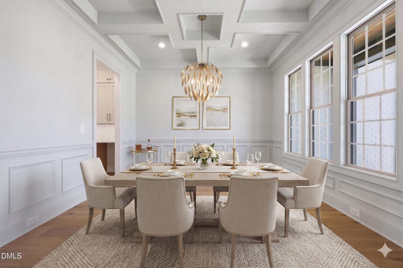 A bright, modern dining room with a light wooden table set for six, cream upholstered chairs, a floral centerpiece, abstract wall art, a gold chandelier, large windows, and white paneled walls.