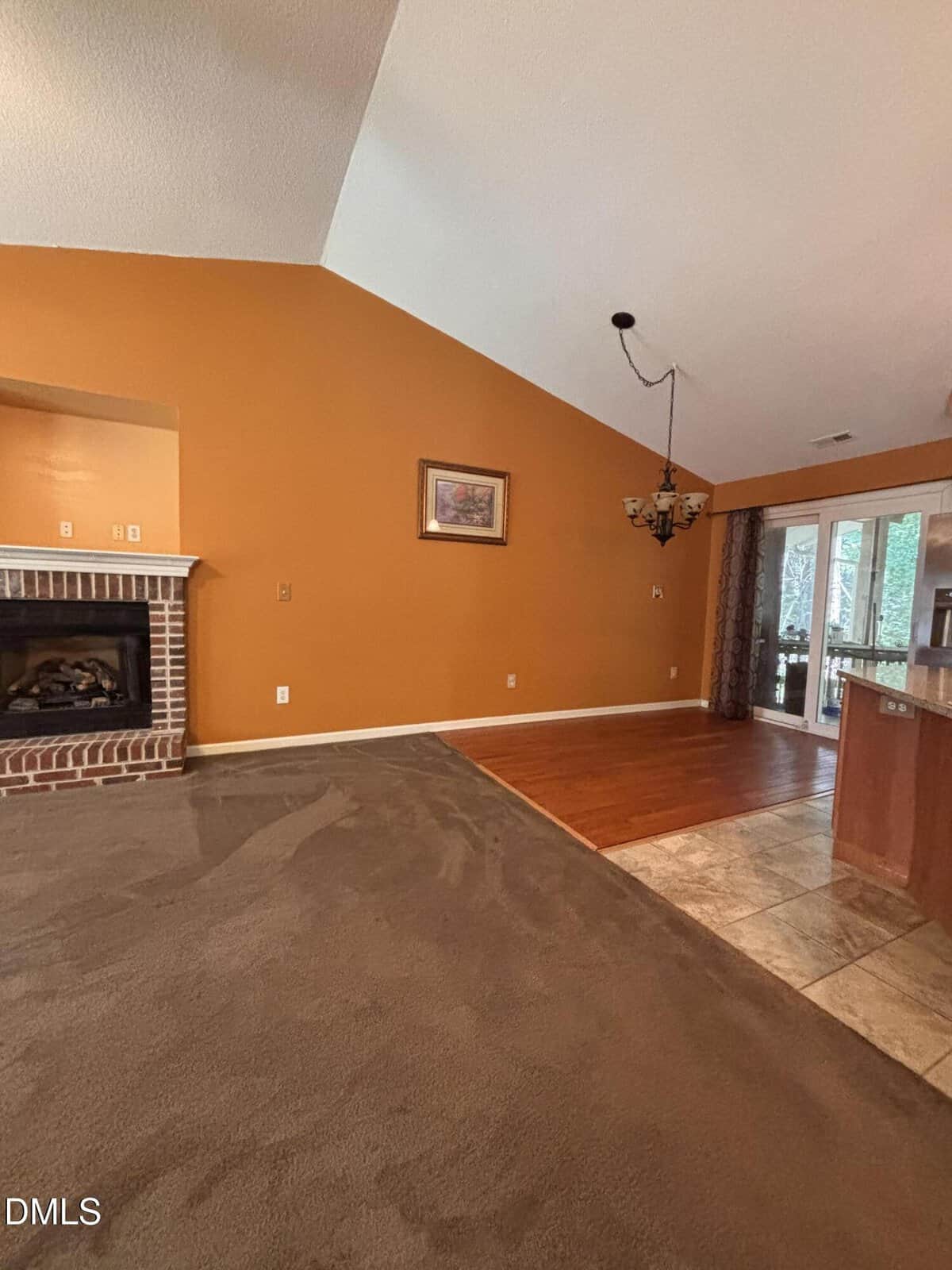 Living room with brown carpet, orange walls, a brick fireplace, hanging light fixture, and framed art. Adjacent area has wood and tile flooring, large windows with curtains, and a view of a patio door.
