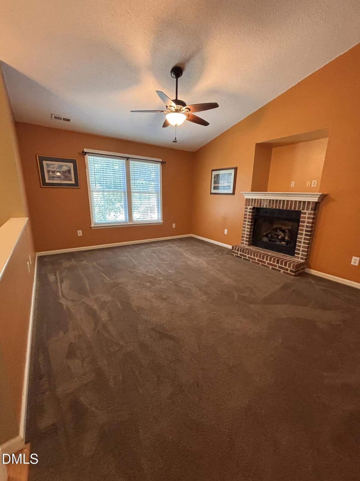 A spacious living room with burnt orange walls, a ceiling fan, large windows with blinds, framed artwork, and a brick fireplace with a niche above it. The carpet is dark beige or brown.