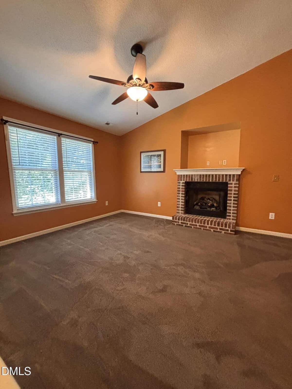 Empty room with orange walls, brown carpet, a ceiling fan, two windows with blinds, a fireplace with a brick mantel, and a framed picture on the wall next to the fireplace.