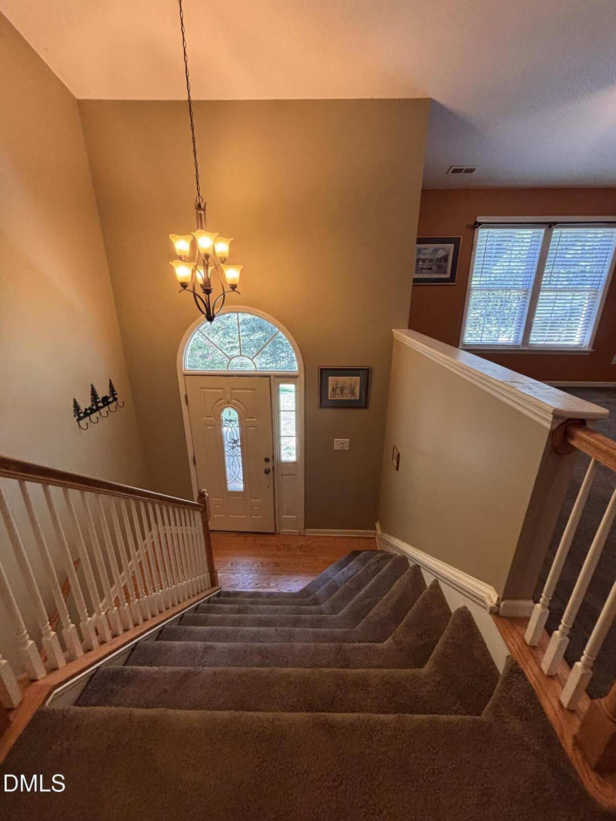 View from the top of a carpeted staircase leading down to a front door with an arched window, a chandelier above, beige walls, white trim, and framed pictures on the walls. Sunlight is coming through nearby windows.