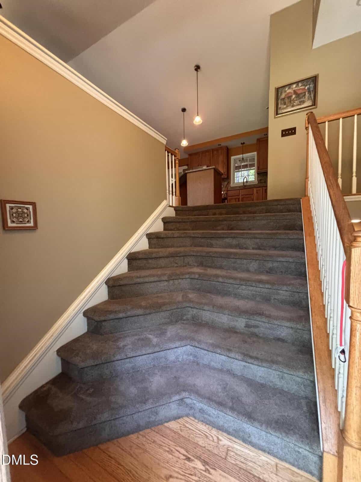 Carpeted staircase with a wooden handrail leads up to a kitchen area with hanging lights. Beige walls feature framed art, and hardwood flooring is at the base of the stairs.