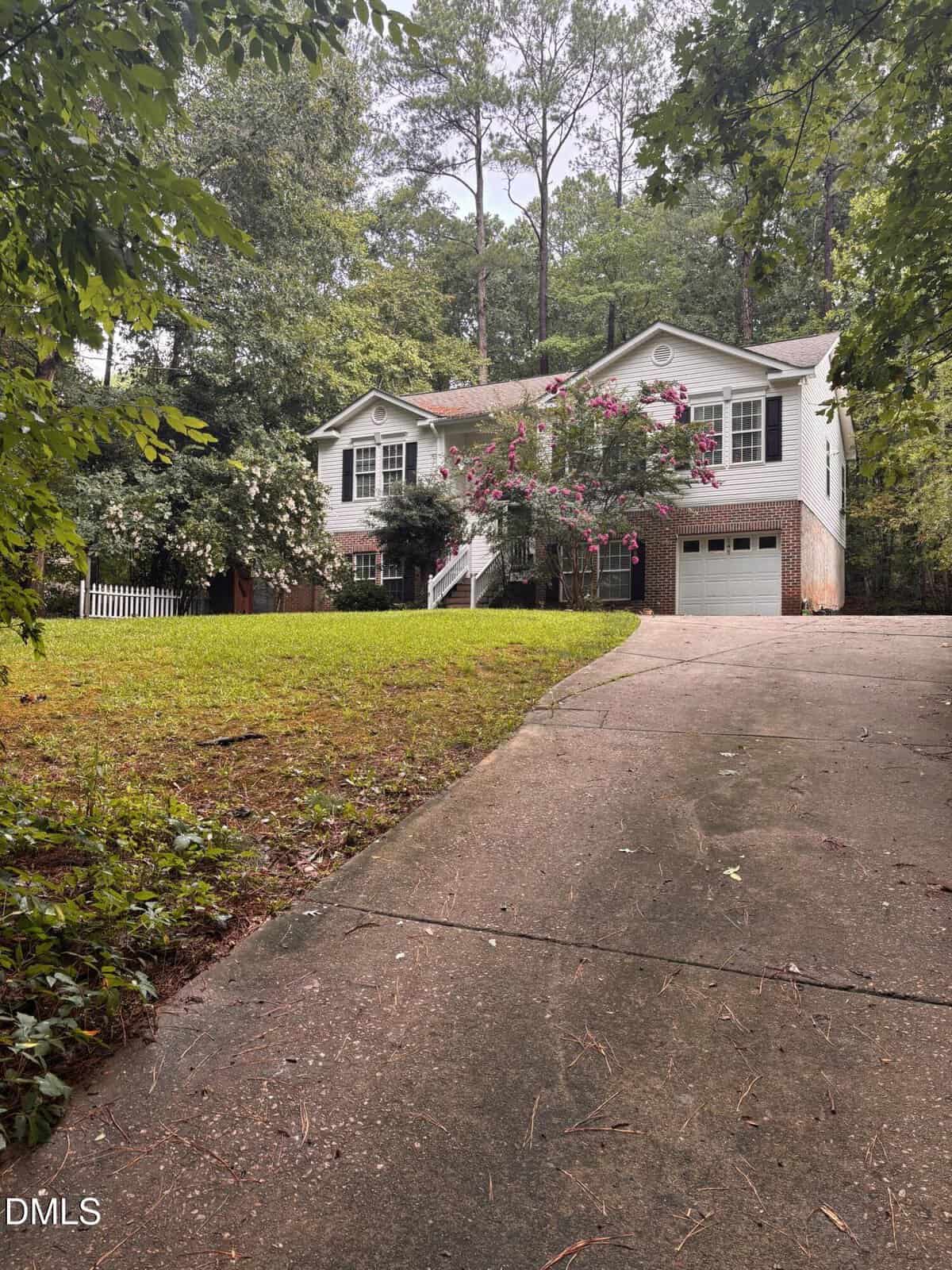 A two-story white house with a garage, front porch, and blooming tree, set among tall trees. A curved driveway leads up to the house through a grassy yard.