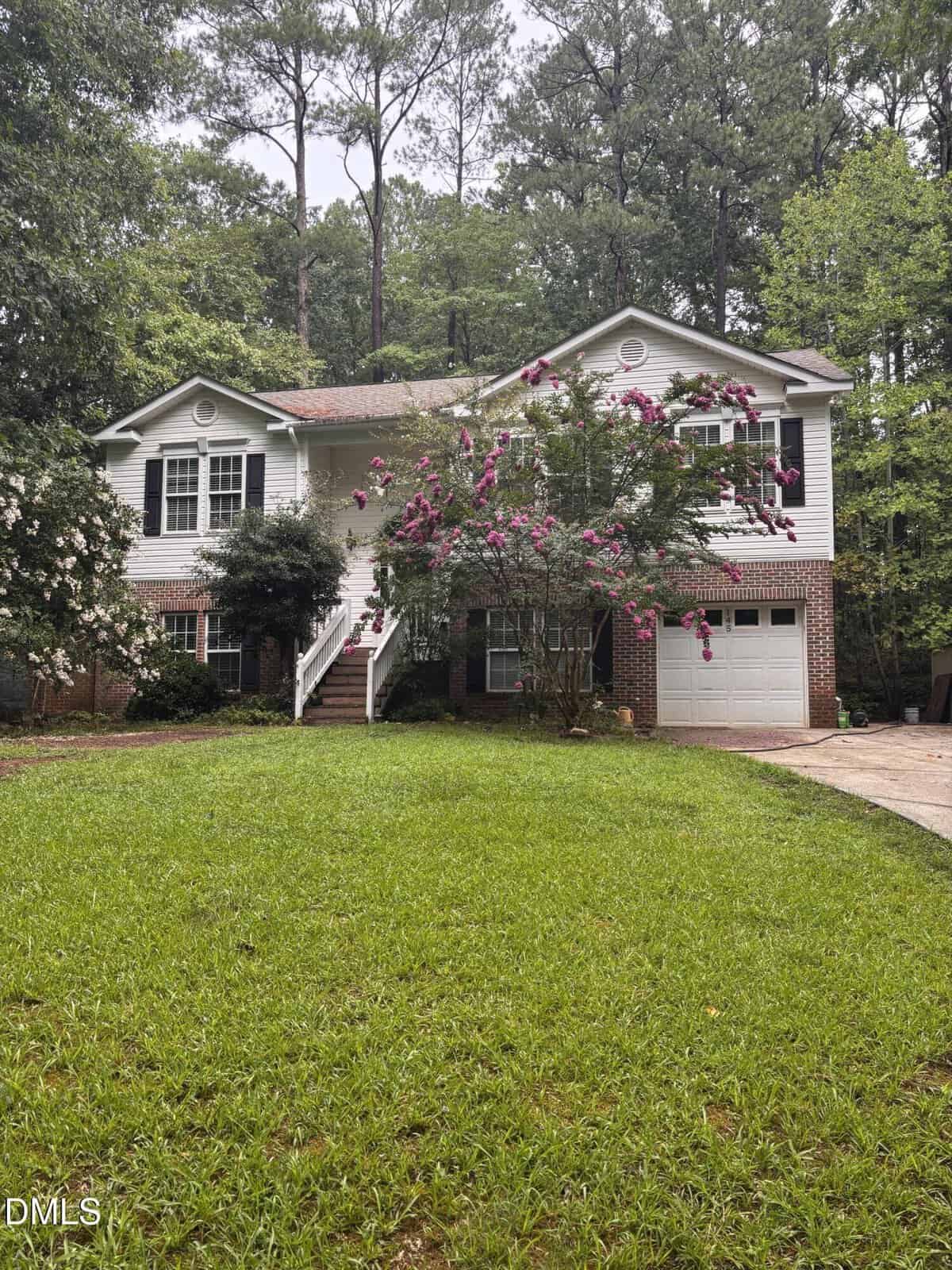 A two-story house with white siding, brick accents, and a garage located at 145 Huron Drive in Louisburg. A tree with pink flowers stands by the front steps. Lush green grass covers the yard, with tall trees and dense foliage in the background.