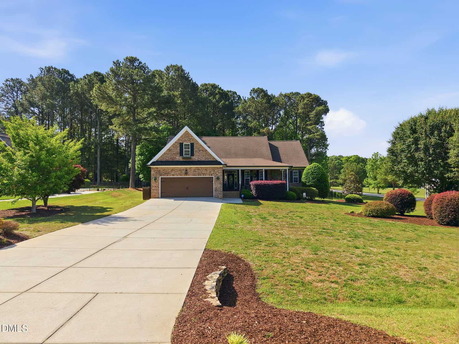 A single-story house with a gabled roof, brick and stone exterior, and attached garage sits at the end of a long concrete driveway, surrounded by a large, neatly landscaped lawn and trees.