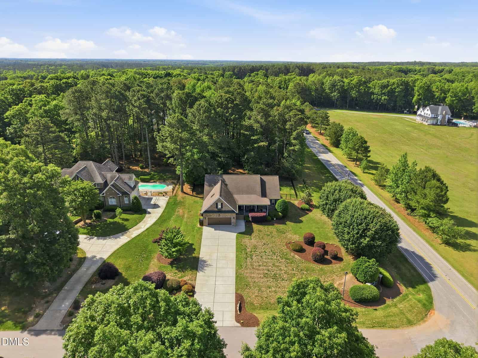 Aerial view of two large houses with driveways, one with a swimming pool, surrounded by trees and greenery, next to a road with fields and forest in the background under a partly cloudy sky.