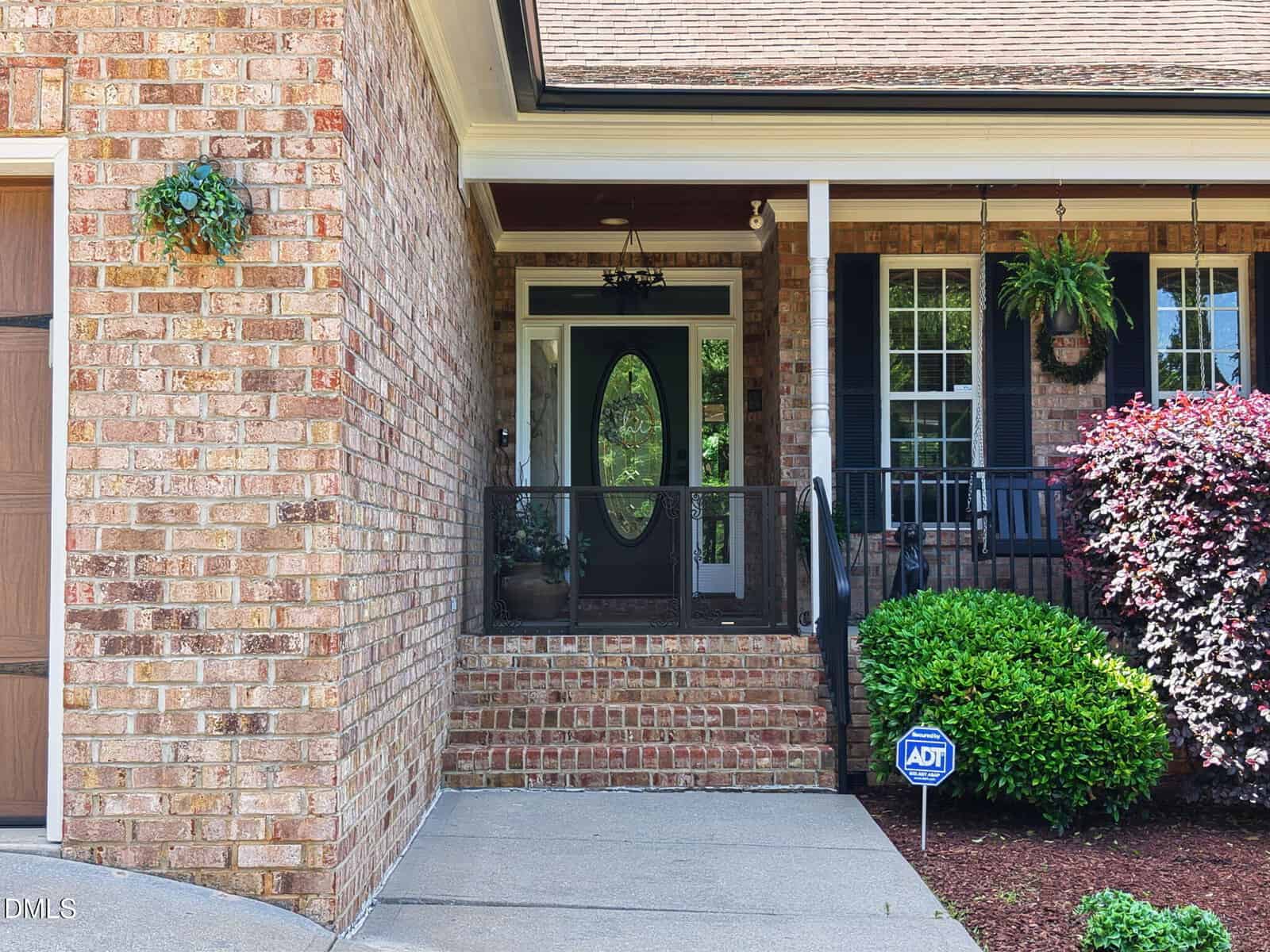 A brick house with black front door, white trim, and a covered porch. Green bushes, hanging plants, and an ADT security sign are visible near the steps.