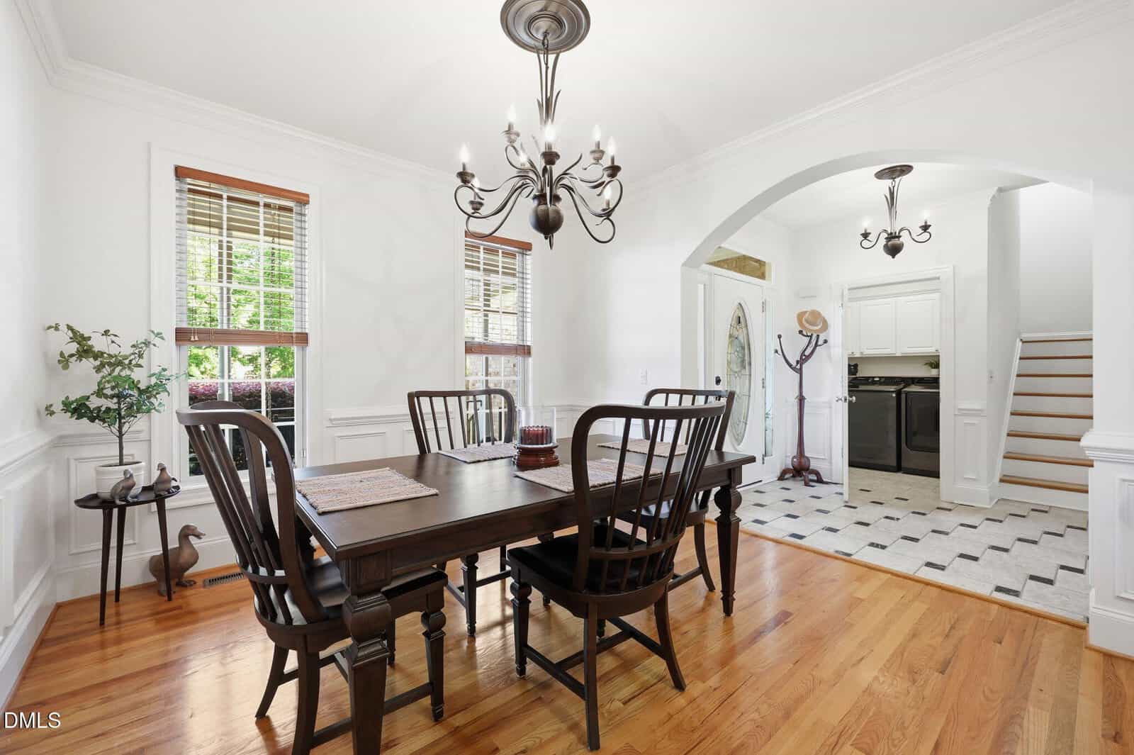 A dining room with a dark wood table and chairs, hardwood floor, chandelier, white walls, windows, and an archway leading to a kitchen and staircase. A coat rack and potted plant are also visible.