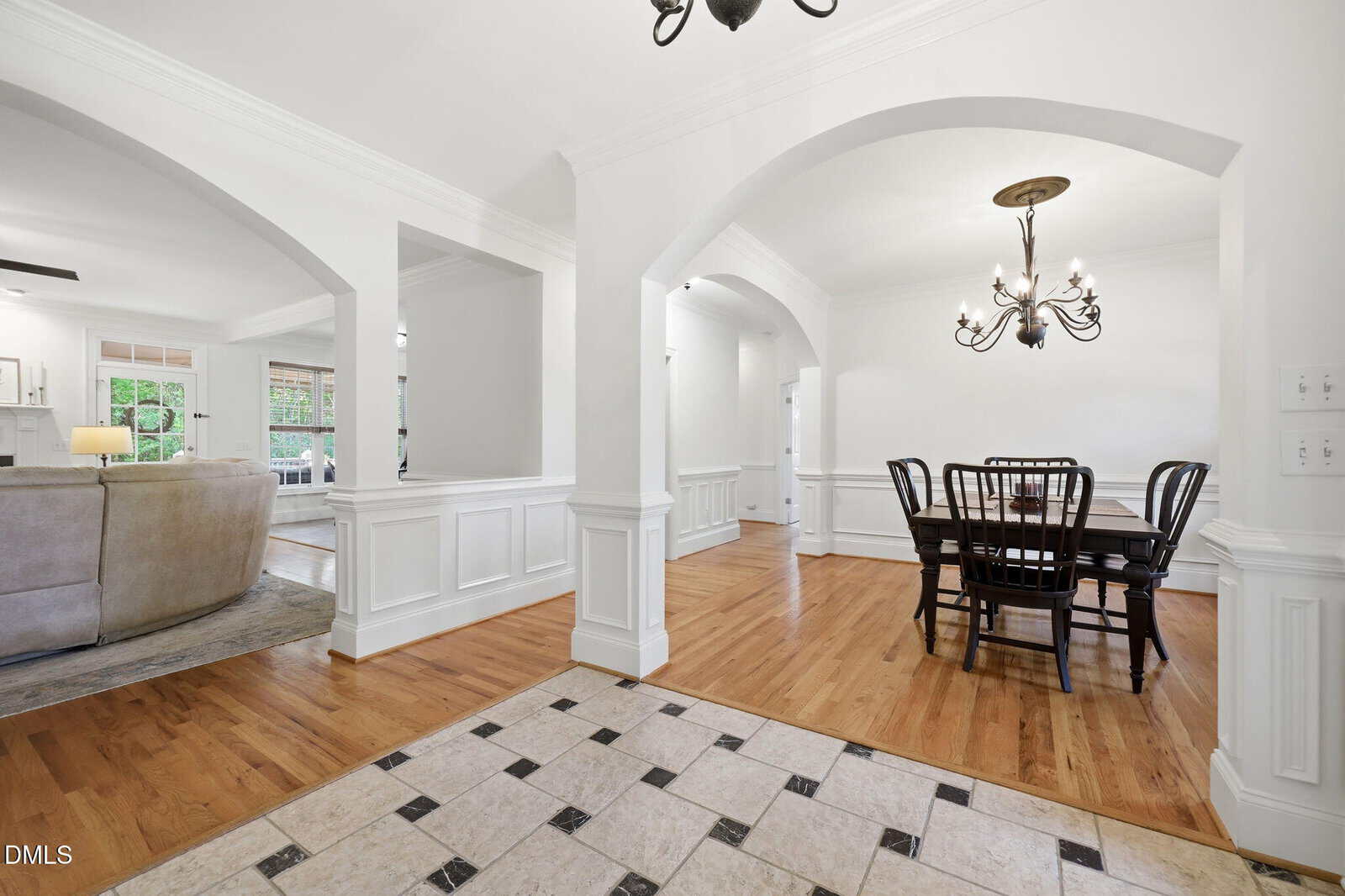 A bright open-plan dining room with wooden floors, a black dining table and chairs, white walls, arched doorways, and a chandelier. Part of a living room and tile flooring are also visible.