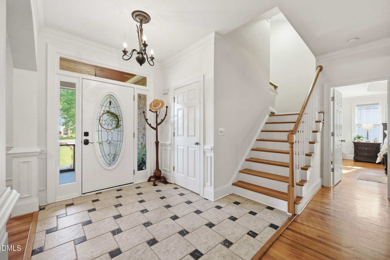 Bright entryway with tiled floor, chandelier, white walls, front door with oval glass, coat rack with hats, wooden staircase with white railing, and hallway leading to a sunlit bedroom.