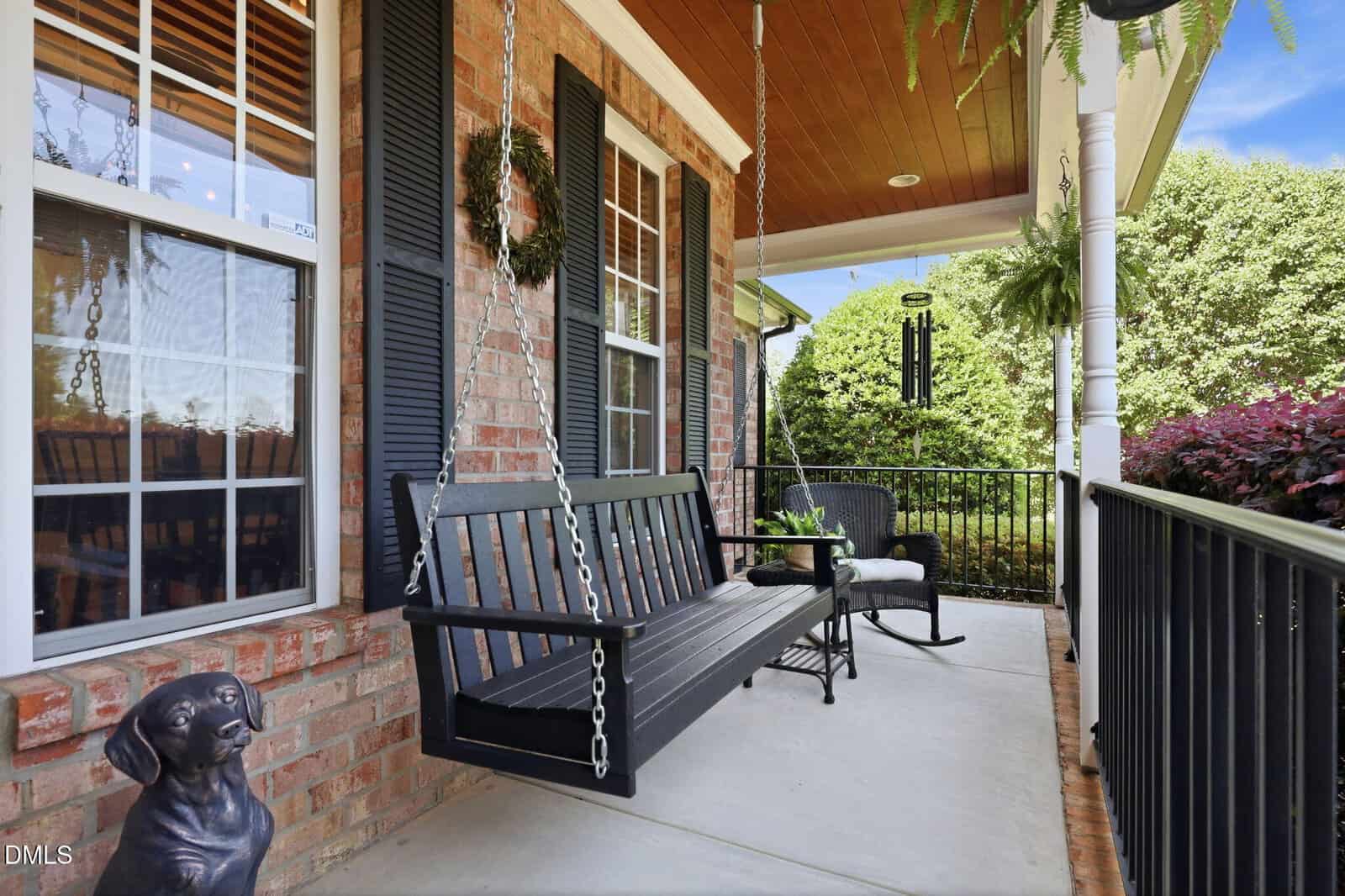 A cozy front porch with a black porch swing, a rocking chair, potted plants, a wind chime, and a dog statue. The porch has brick walls, black shutters, and a wooden ceiling with hanging ferns.