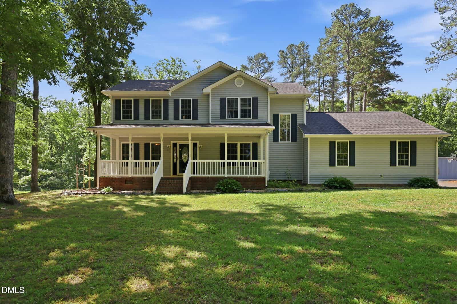 Two-story house with gray siding, dark shutters, and a large front porch, surrounded by green grass, trees, and a clear blue sky. A single-story section extends to the right.