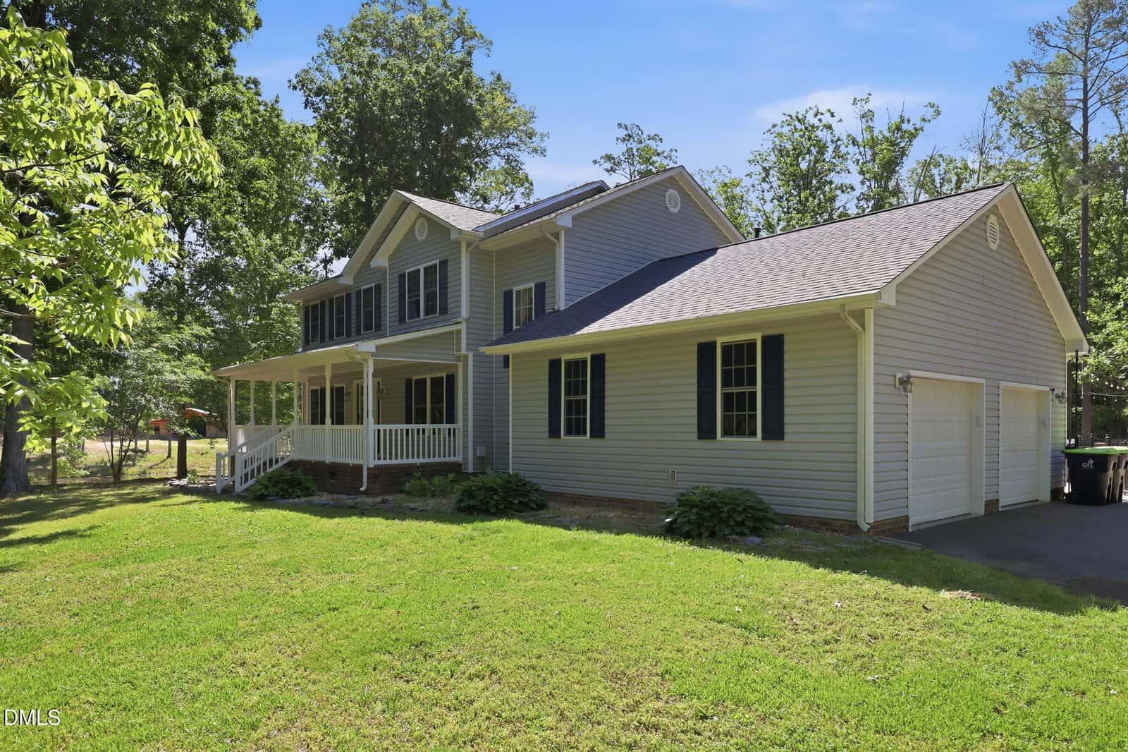 Two-story house with light gray siding, white trim, a covered front porch, and an attached two-car garage, surrounded by green grass, trees, and a paved driveway.