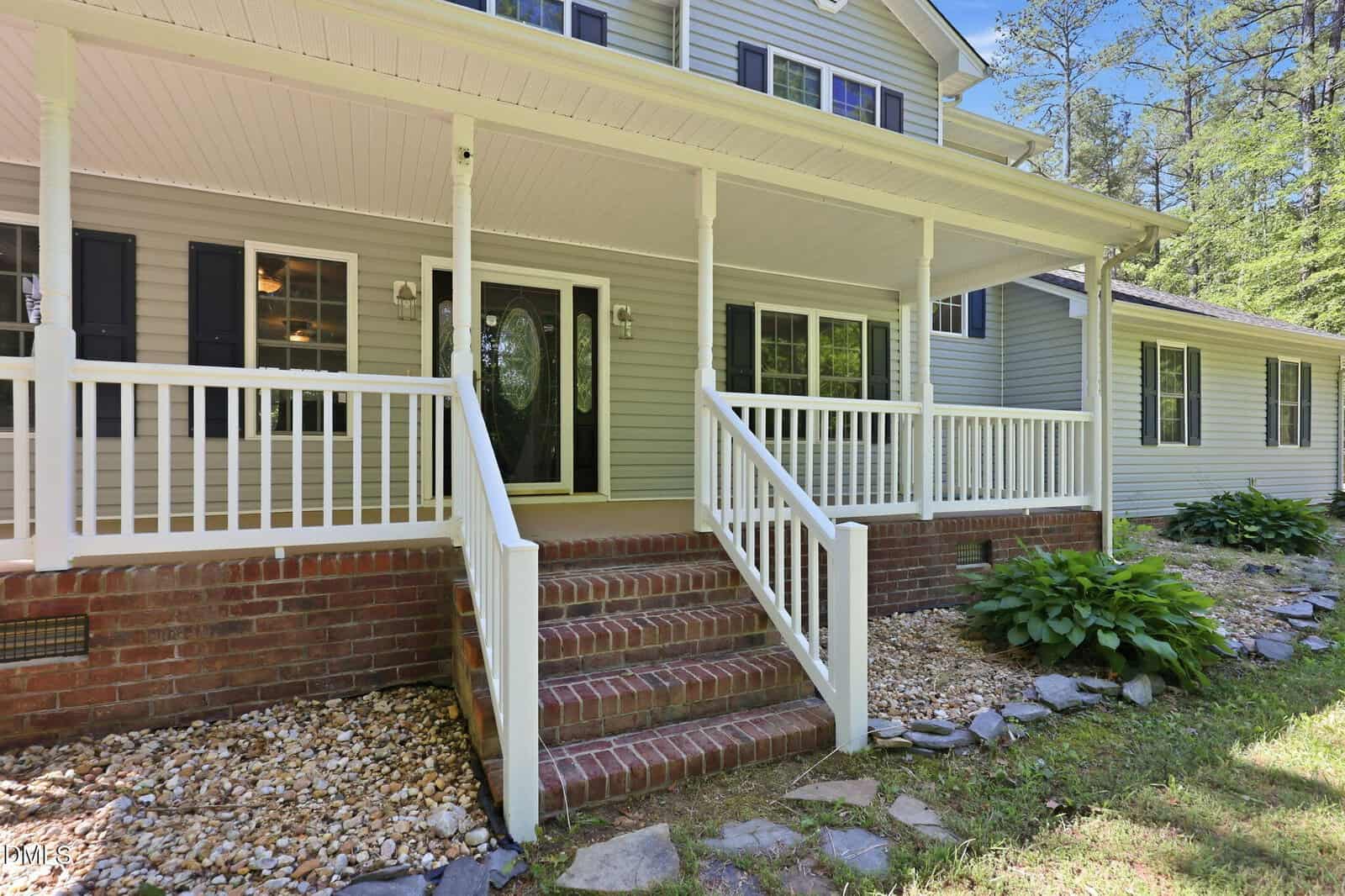 A front view of a house with light gray siding, black shutters, and a covered porch featuring white railings and steps leading up from a brick walkway, surrounded by landscaping with rocks and greenery.