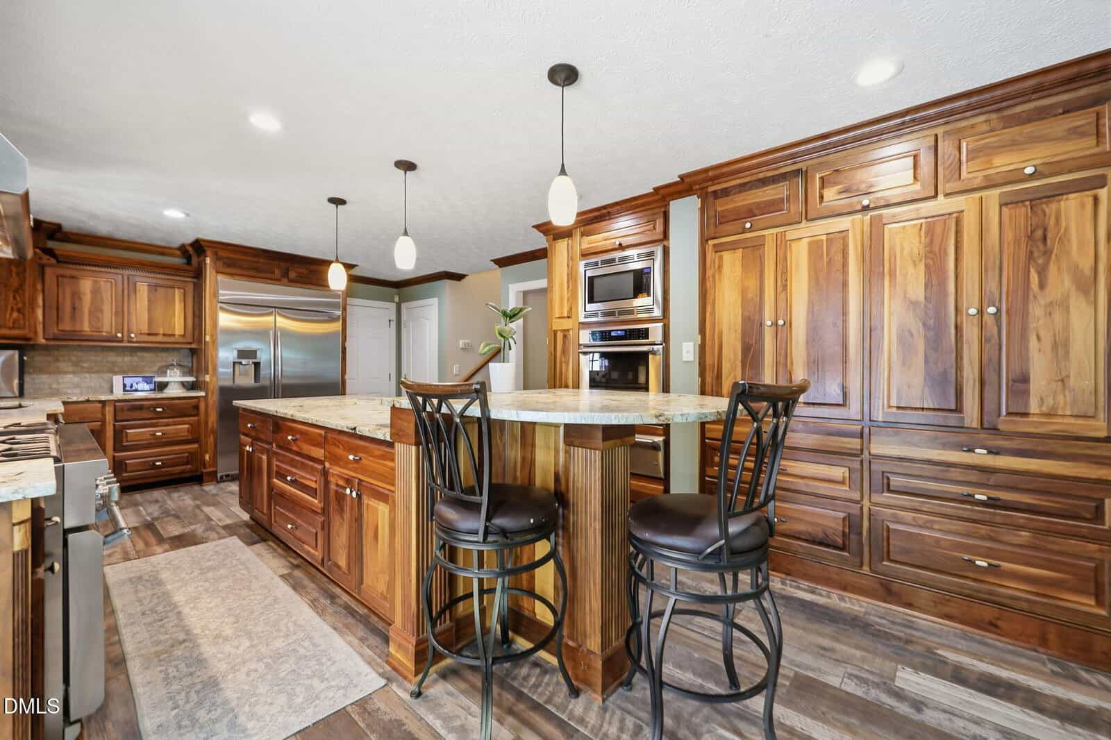 Spacious kitchen with wood cabinetry, stainless steel appliances, a large island with granite countertops, and two black metal barstools. Pendant lights hang from the ceiling, and the floor is wood-patterned.