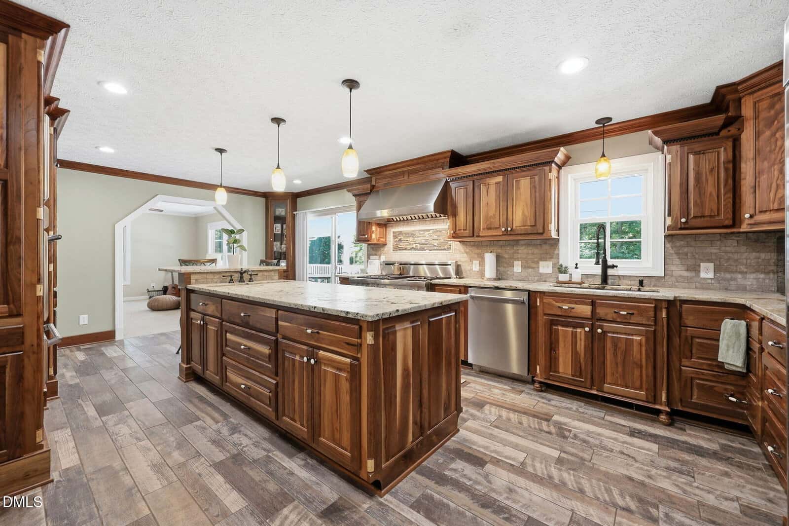 Spacious kitchen with wooden cabinets, granite countertops, a central island, stainless steel appliances, pendant lights, and large windows letting in natural light. The floor features wide, rustic wood planks.
