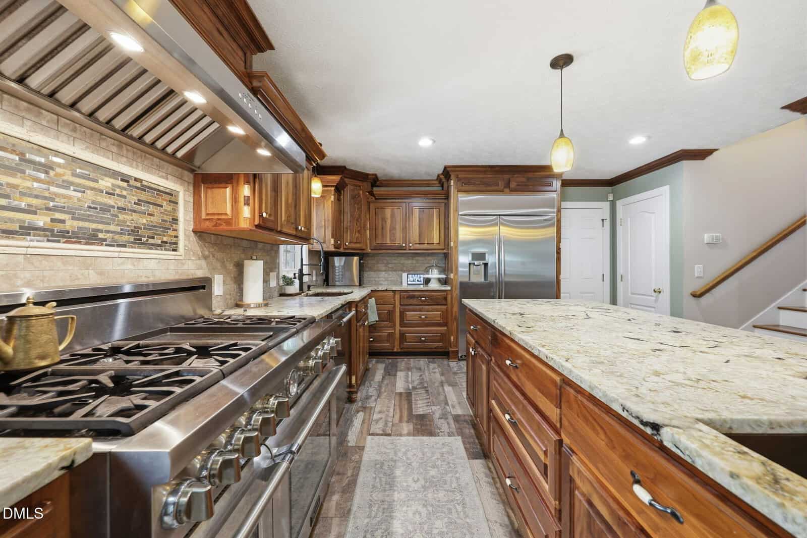 Spacious kitchen with granite countertops, stainless steel appliances, wooden cabinets, a large stove, double-door fridge, pendant lights, and a tile backsplash. Stairs and a closed door are visible in the background.