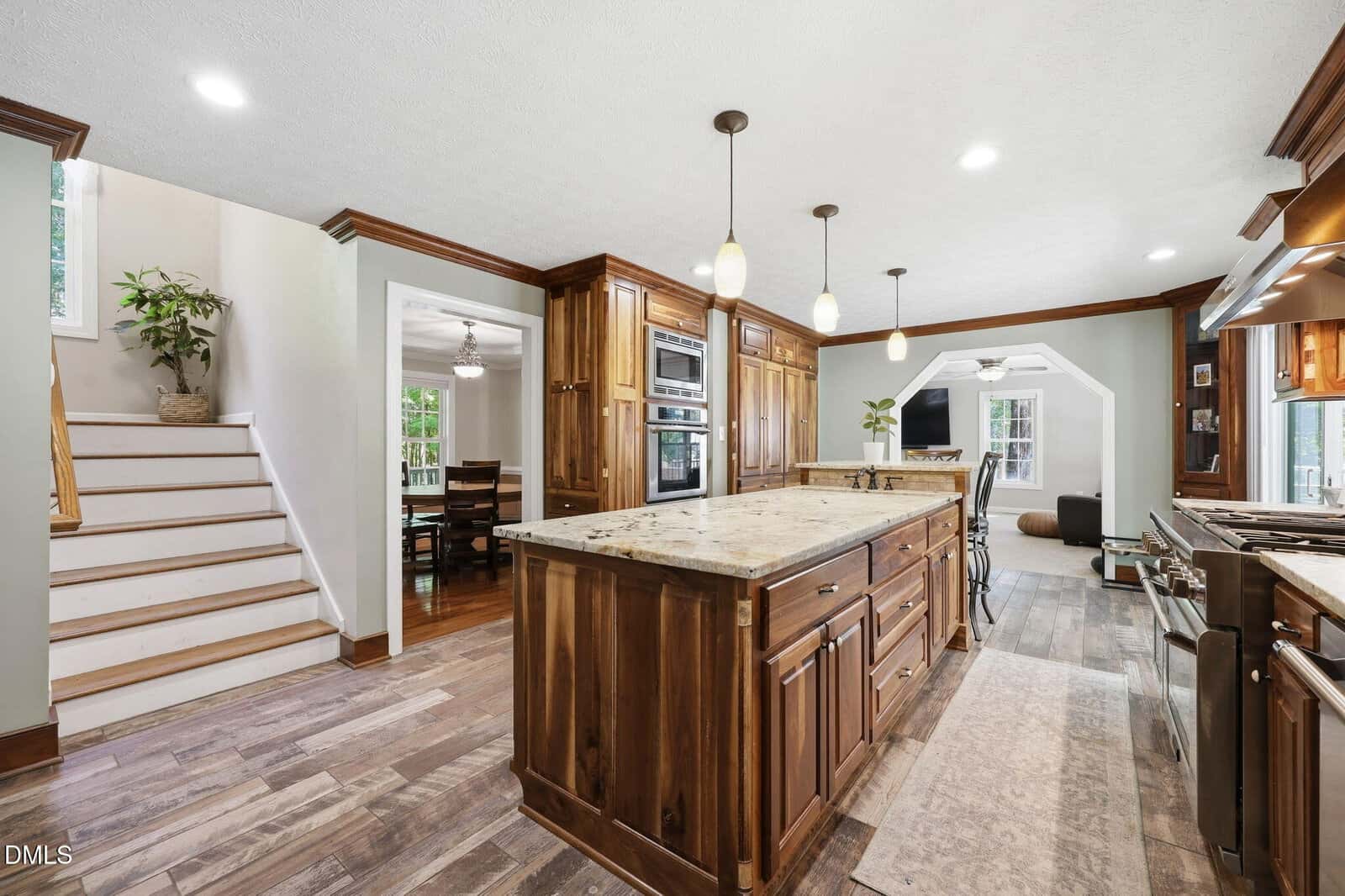 Spacious kitchen with wood cabinets, center island, pendant lights, and stainless steel appliances. Wooden stairs and dining area are visible. Large windows provide natural light.