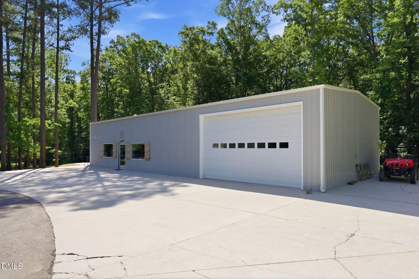 A large metal building with a white garage door and glass entry doors sits on a wide concrete driveway, surrounded by tall trees and greenery on a sunny day.