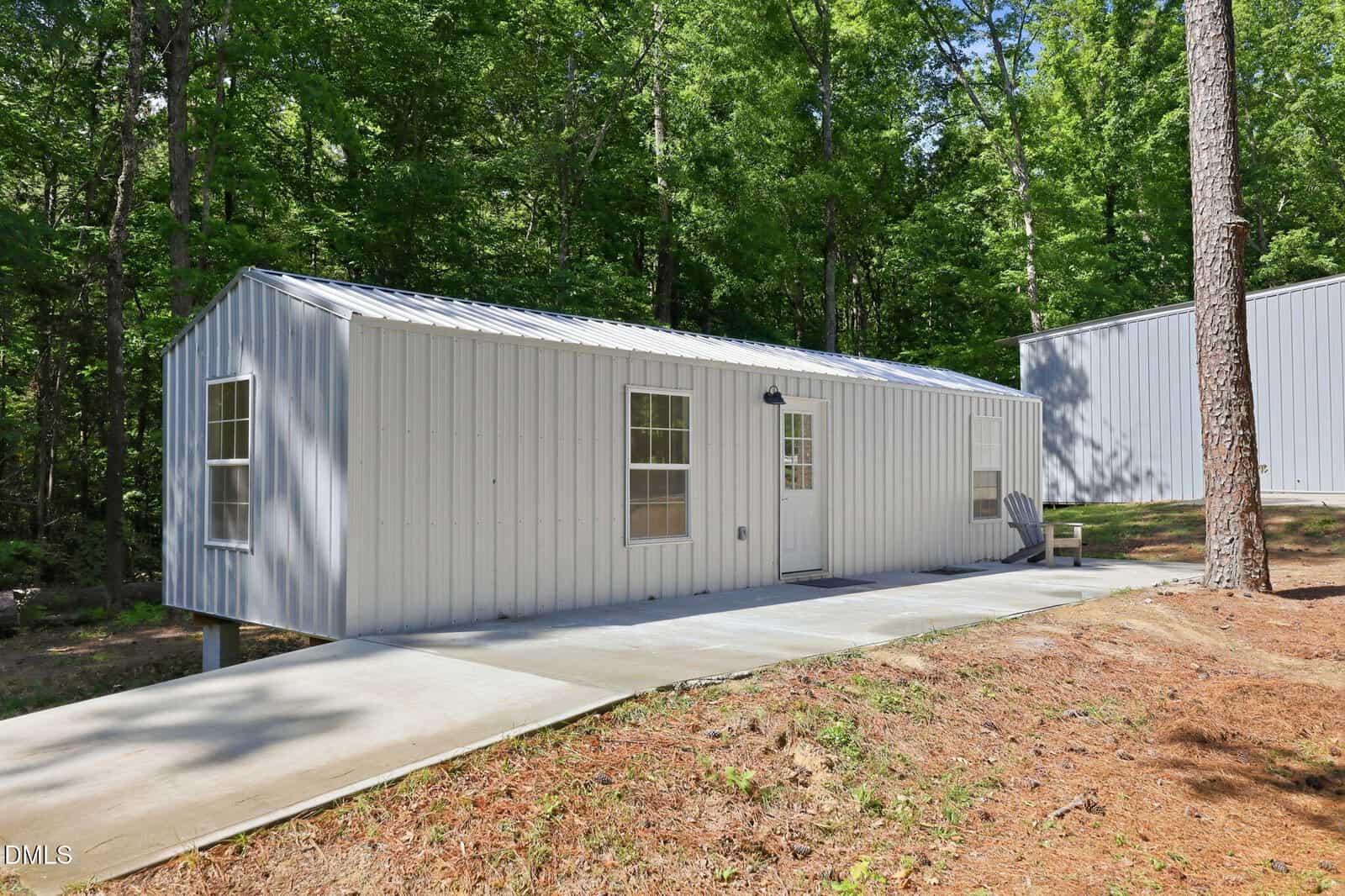 A small white metal building with a sloped roof, three windows, and a door sits on a concrete path surrounded by trees and greenery. The structure is elevated slightly above the sloping ground.