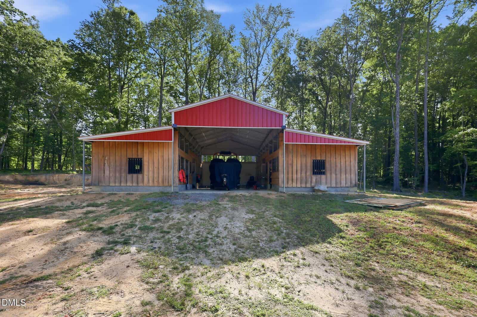 A red and wood barn with open central doors, revealing a horse inside. The structure is surrounded by green grass and tall trees under a clear blue sky.