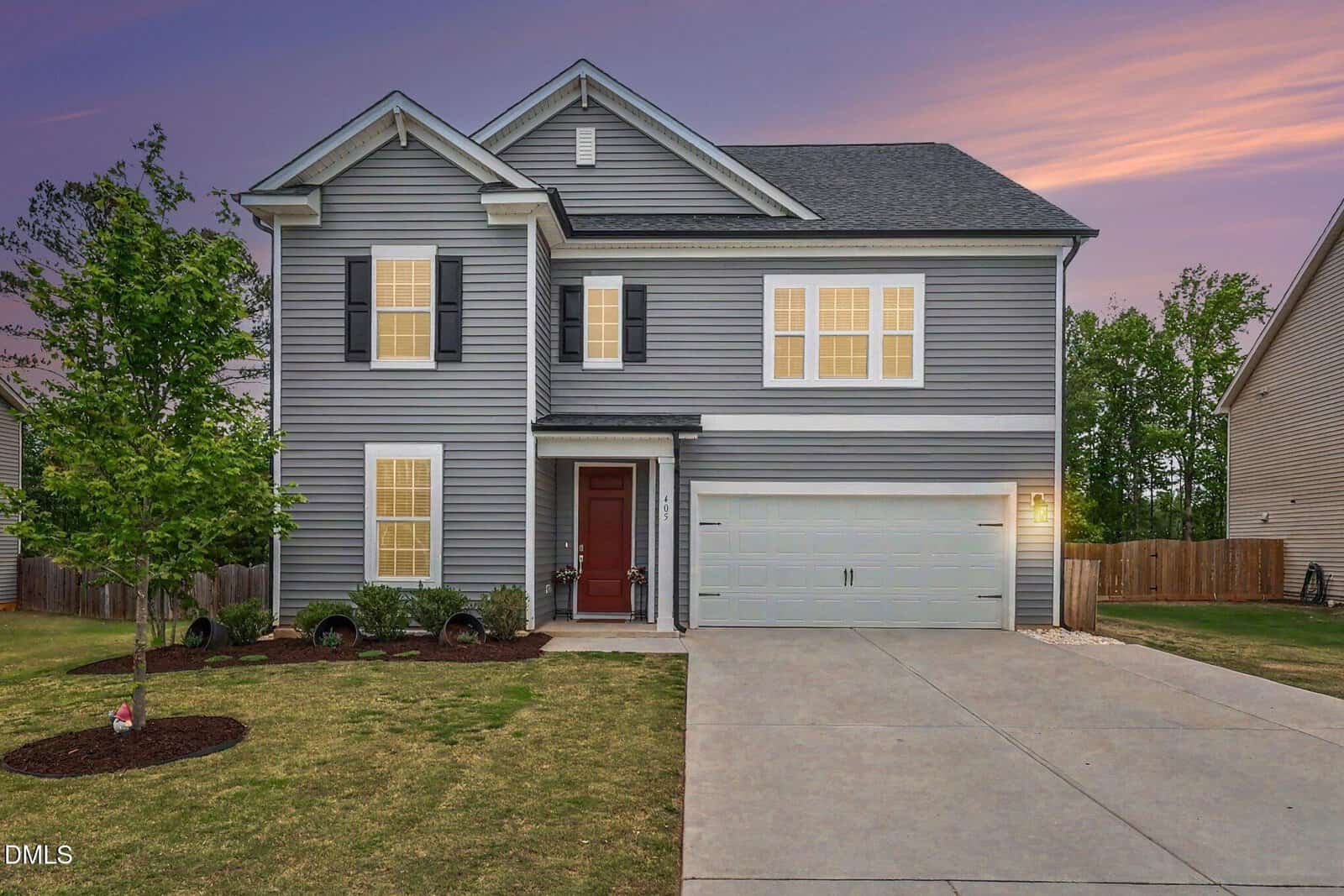 Two-story gray house with white trim, black shutters, and a red front door. The home has a two-car garage, a well-kept lawn, a small tree, and bushes along the front. The sky is purple and orange at sunset.