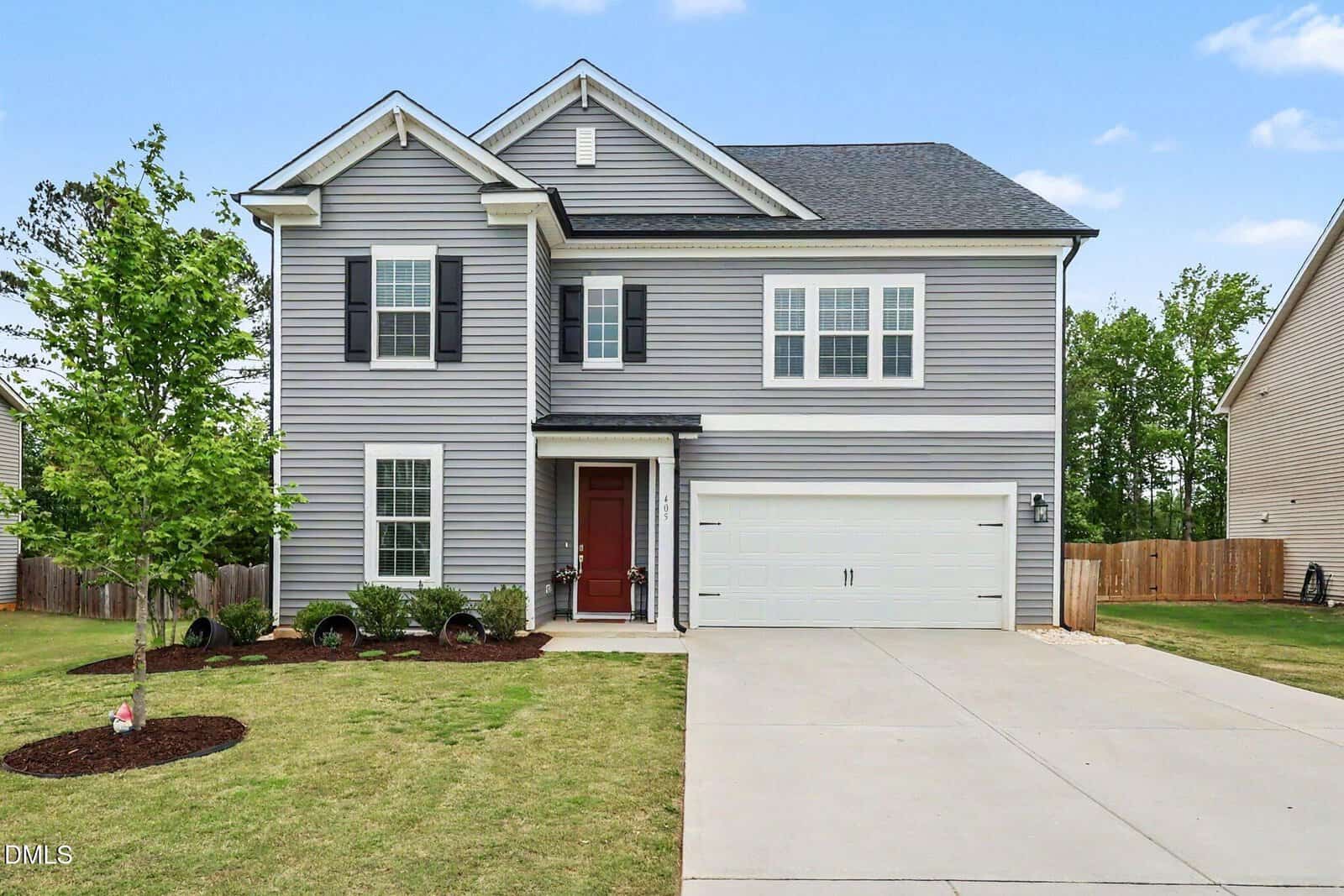 Two-story gray house with white trim, black shutters, and a red front door. Attached two-car garage, neatly mowed lawn, young tree, and shrub landscaping in the front yard. Clear blue sky in the background.