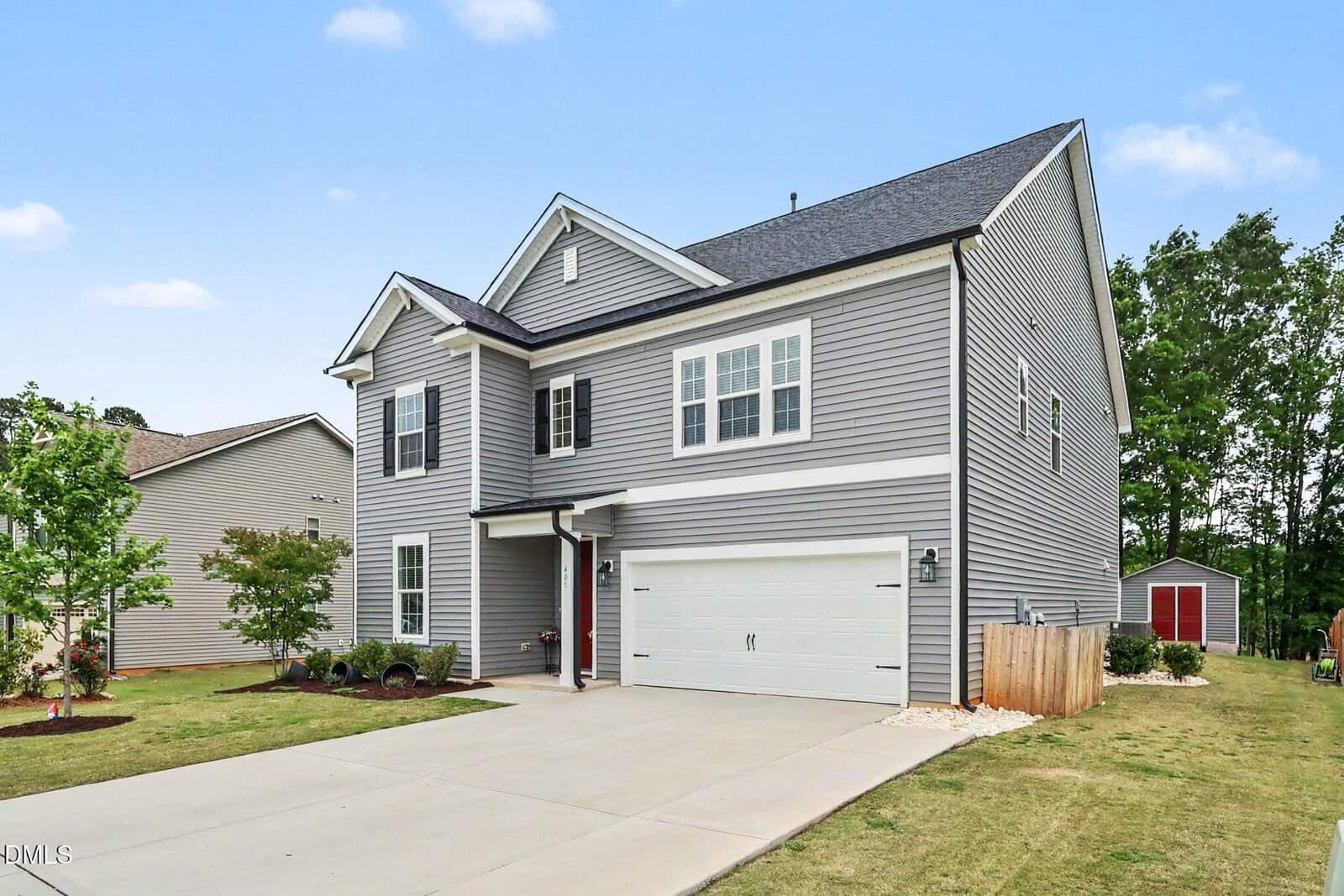 A modern two-story gray house with white trim, two-car garage, driveway, manicured lawn, and trees. The home has multiple windows and is set against a blue sky with scattered clouds.