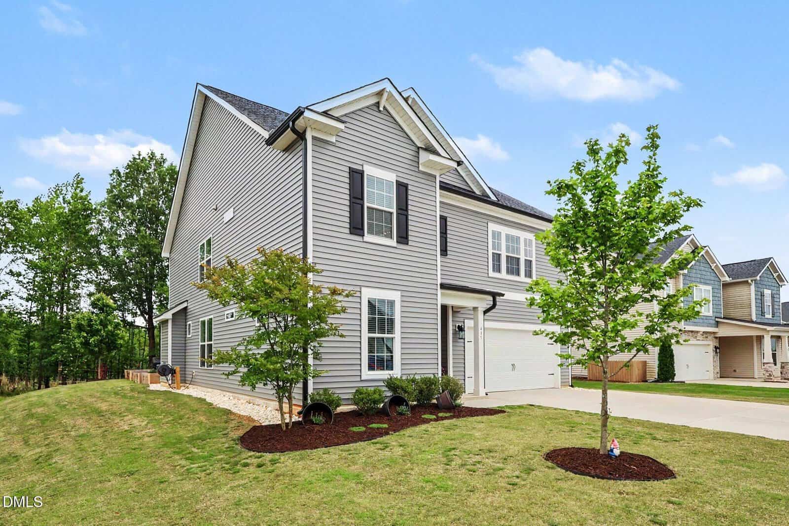 A modern two-story gray house with white trim and black shutters, featuring a front lawn with young trees and shrubs, a double garage, and neighboring homes visible under a blue sky with scattered clouds.