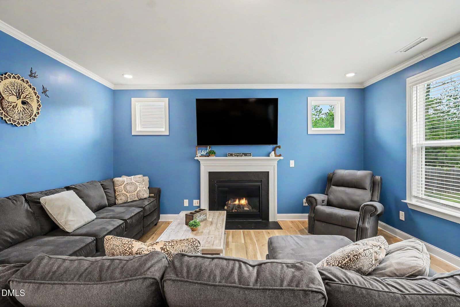 A cozy living room with blue walls, grey sectional sofas, an armchair, a fireplace with a TV above it, and windows letting in natural light. A decorative wall clock hangs on the left.