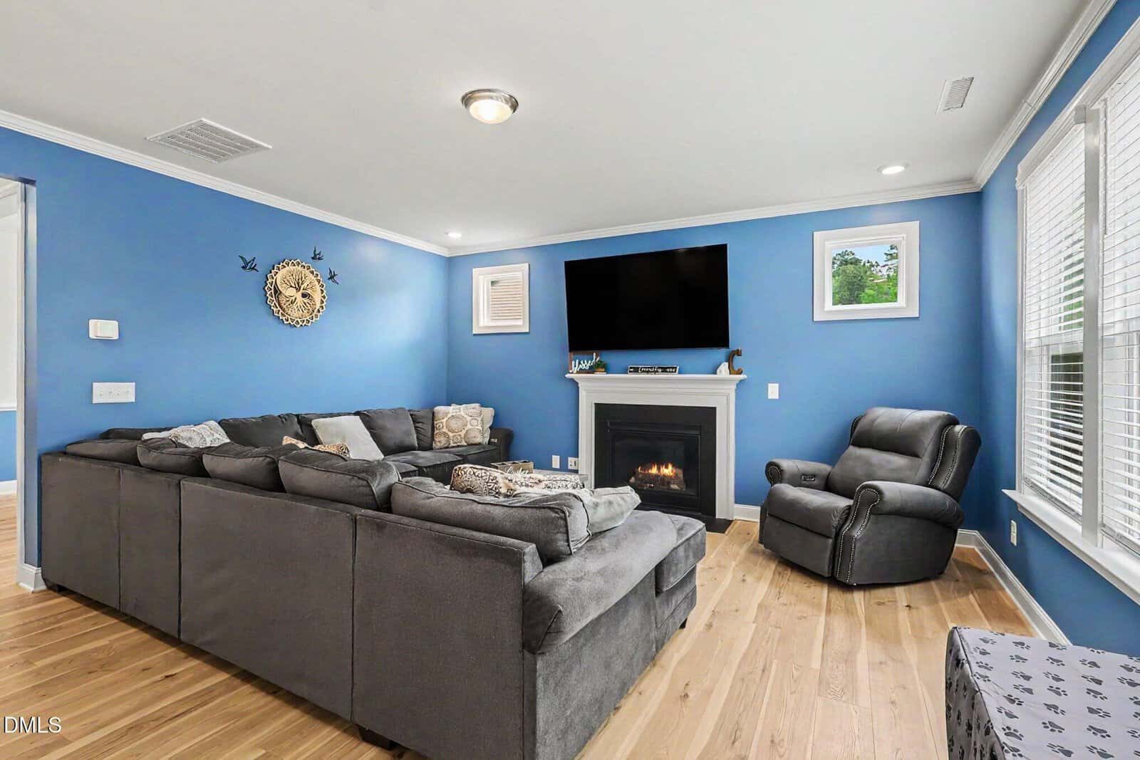 Living room with blue walls, gray sectional sofa, gray armchair, fireplace, wall-mounted TV, hardwood floors, and large windows letting in natural light.