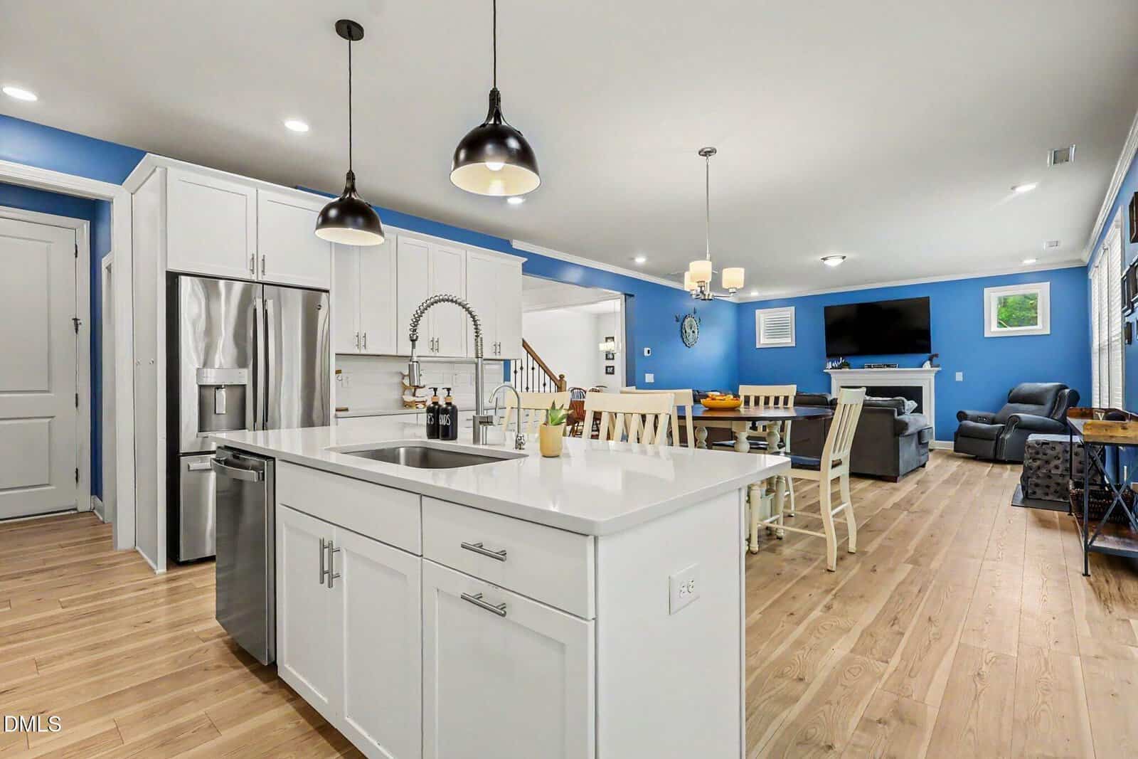 Modern open kitchen with white cabinets, stainless steel appliances, island with sink, wooden floor, blue walls, dining area, and living room with TV and recliners in the background. Black pendant lights hang above the island.