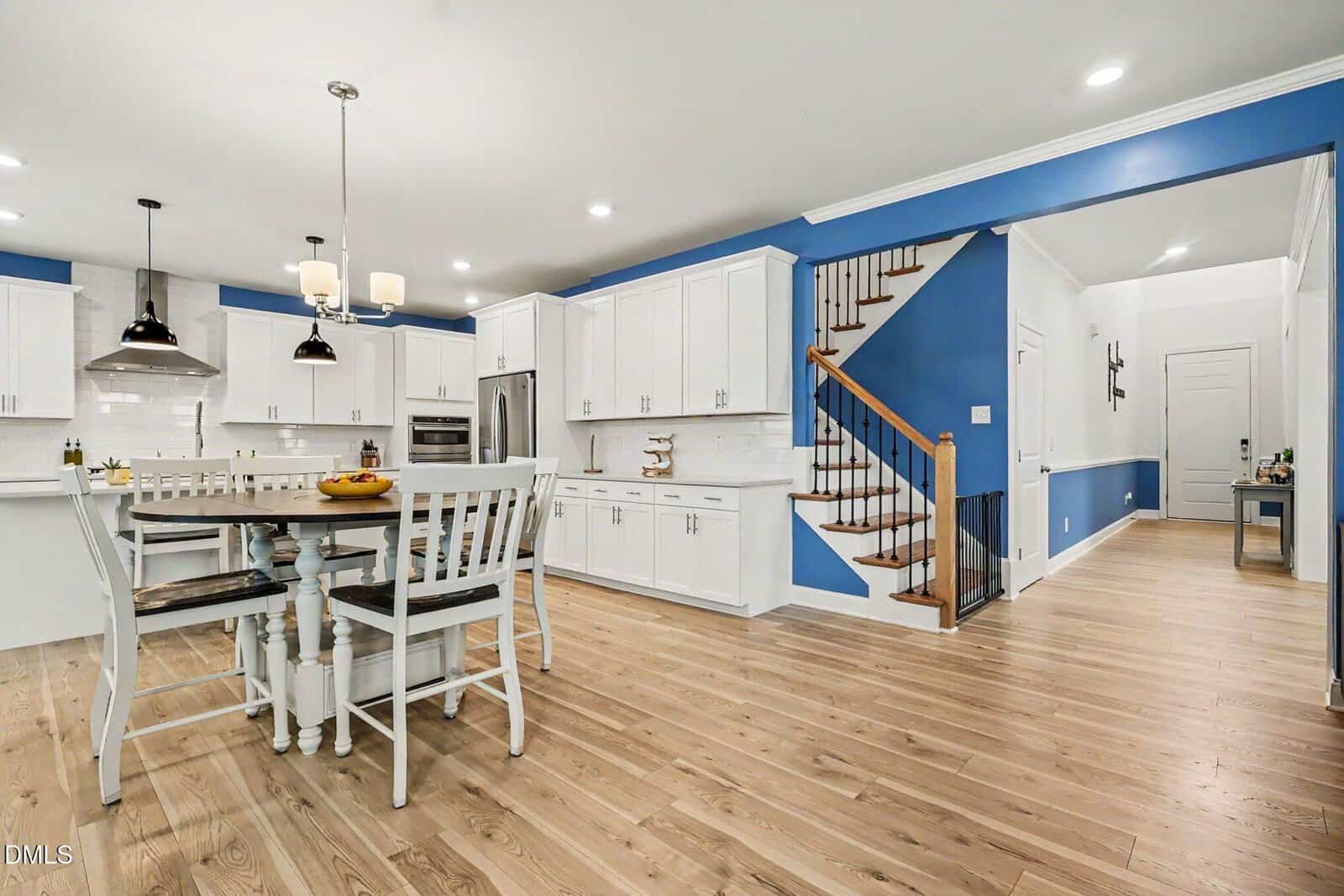 Modern kitchen with white cabinets, stainless steel appliances, a dining table with four chairs, wooden floors, blue accent walls, and a staircase with wooden steps and a black railing.