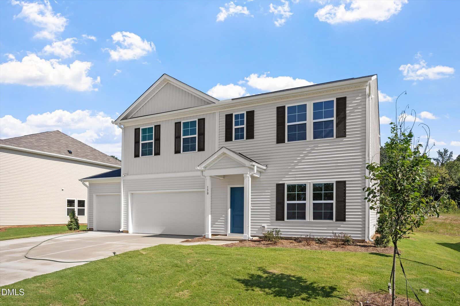 Two-story suburban house with white siding, dark shutters, a blue front door, three-car garage, and neatly cut green lawn under a blue sky with scattered clouds. A young tree is planted in the front yard.