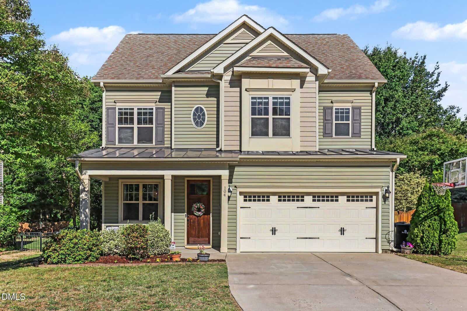 This two-story suburban house at 104 Bonterra Drive, Youngsville, features tan siding, white trim, a double garage, small front porch, manicured shrubs, and a concrete driveway on a green lawn with trees in the background.