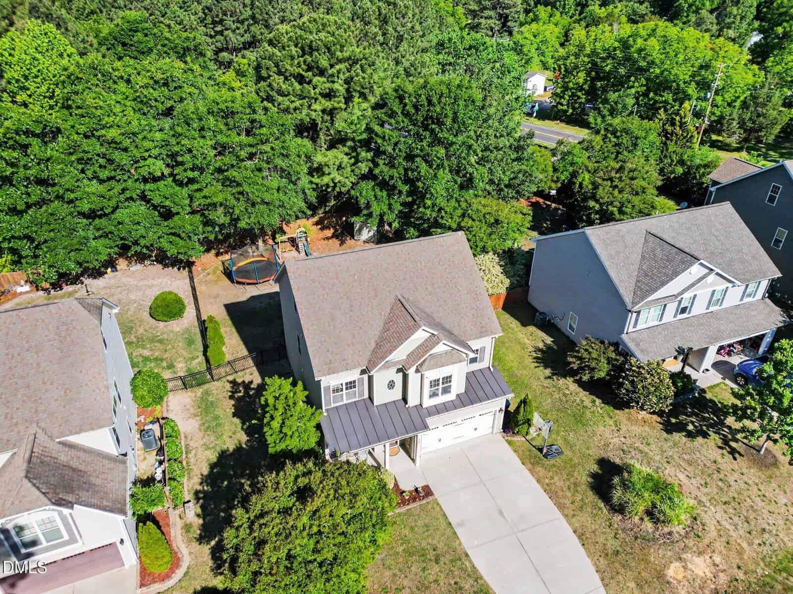 Aerial view of a suburban neighborhood showing three houses with driveways, lawns, and trees. The central house has a fenced backyard with a trampoline and is bordered by greenery and other homes.