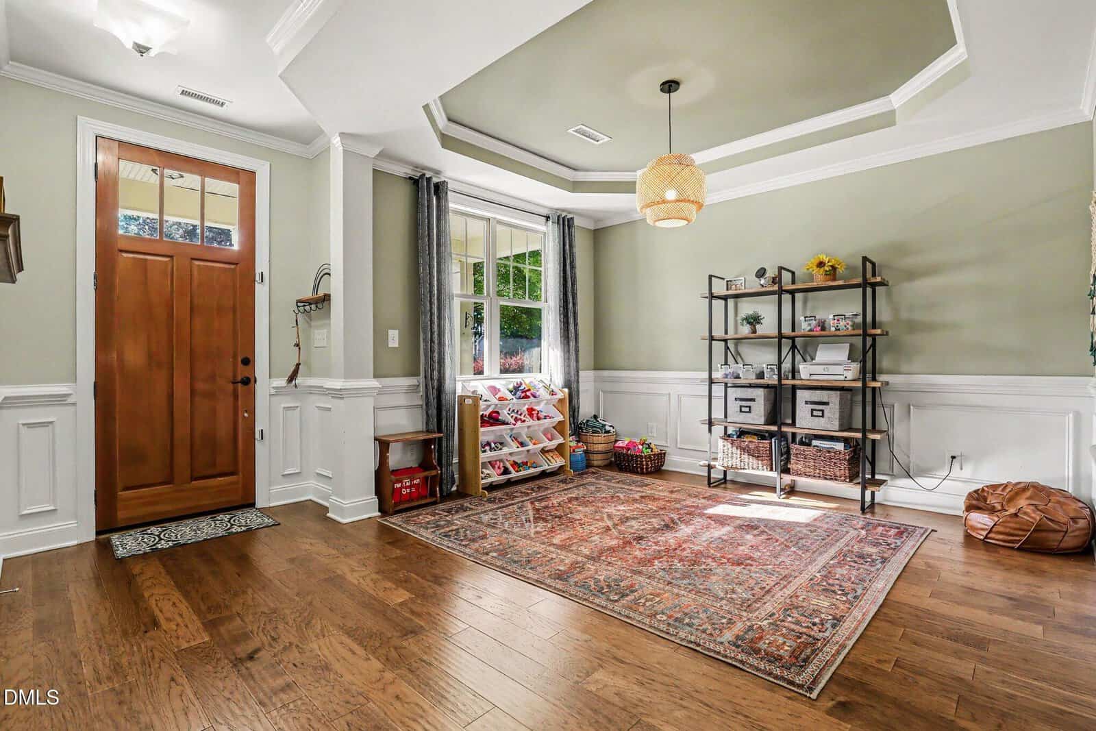 Bright, organized playroom with wood floors, a patterned rug, toy storage by a large window, a metal shelf with baskets and toys, and a wooden front door with transom window. Neutral walls, white trim, and a pendant light overhead.