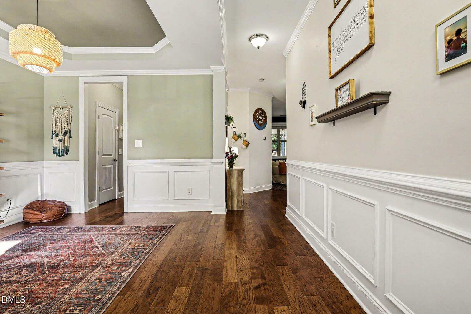 A hallway with white wainscoting, hardwood floors, and beige walls. A patterned rug, hanging light fixture, wall art, and shelves decorate the space. Doorways and natural light are visible in the background.
