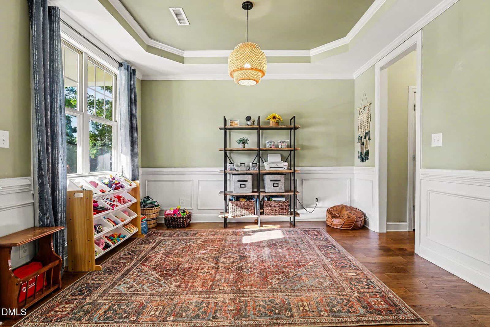 A bright, organized playroom with a large patterned rug, a toy shelf, baskets, a metal bookshelf with bins and toys, wooden floors, green walls, white trim, and a woven pendant light hanging from a tray ceiling.