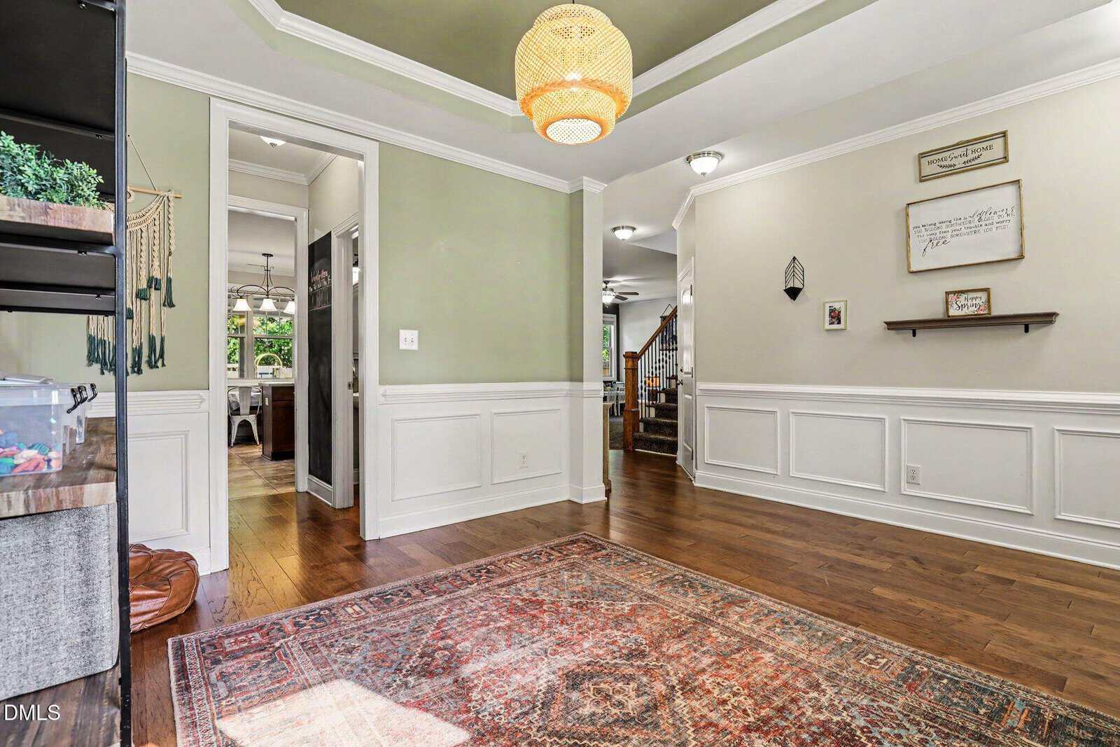 A spacious room with hardwood floors, a patterned rug, green walls, white wainscoting, a tray ceiling with a woven light fixture, and decor on the walls. Open doorways lead to a hallway and another room.