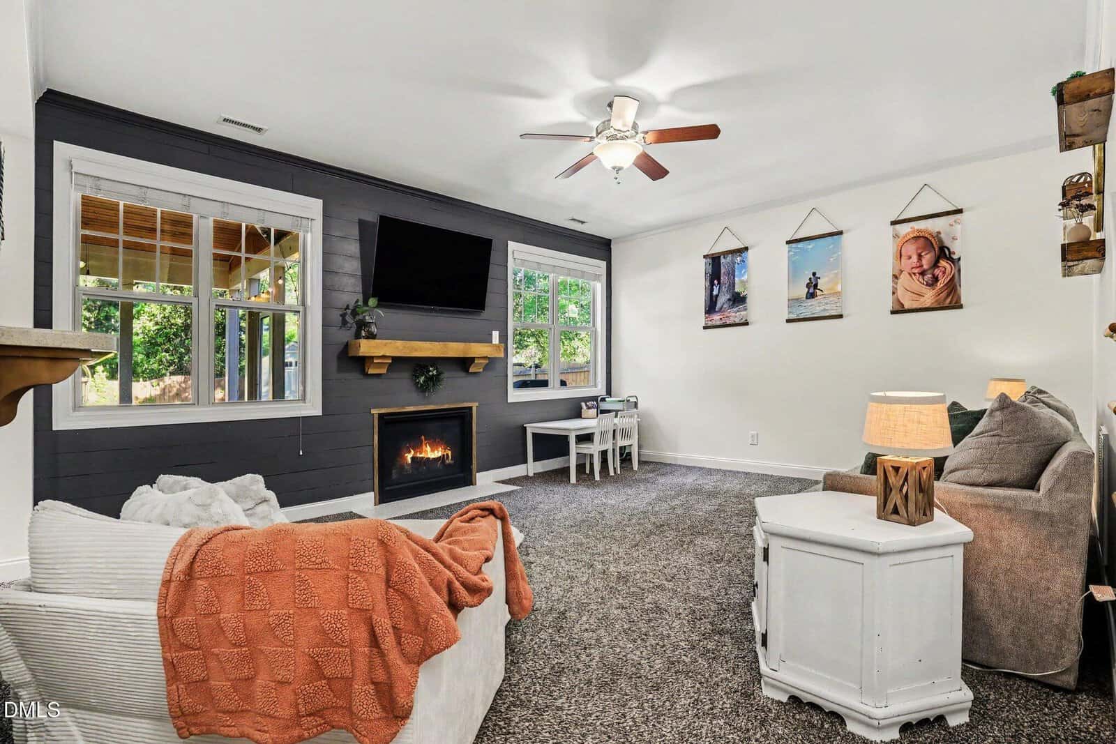 A cozy living room with a gray accent wall, fireplace, mounted TV, white furniture, carpeted floor, and family photos on the wall. A window looks out onto greenery, and a ceiling fan is overhead.