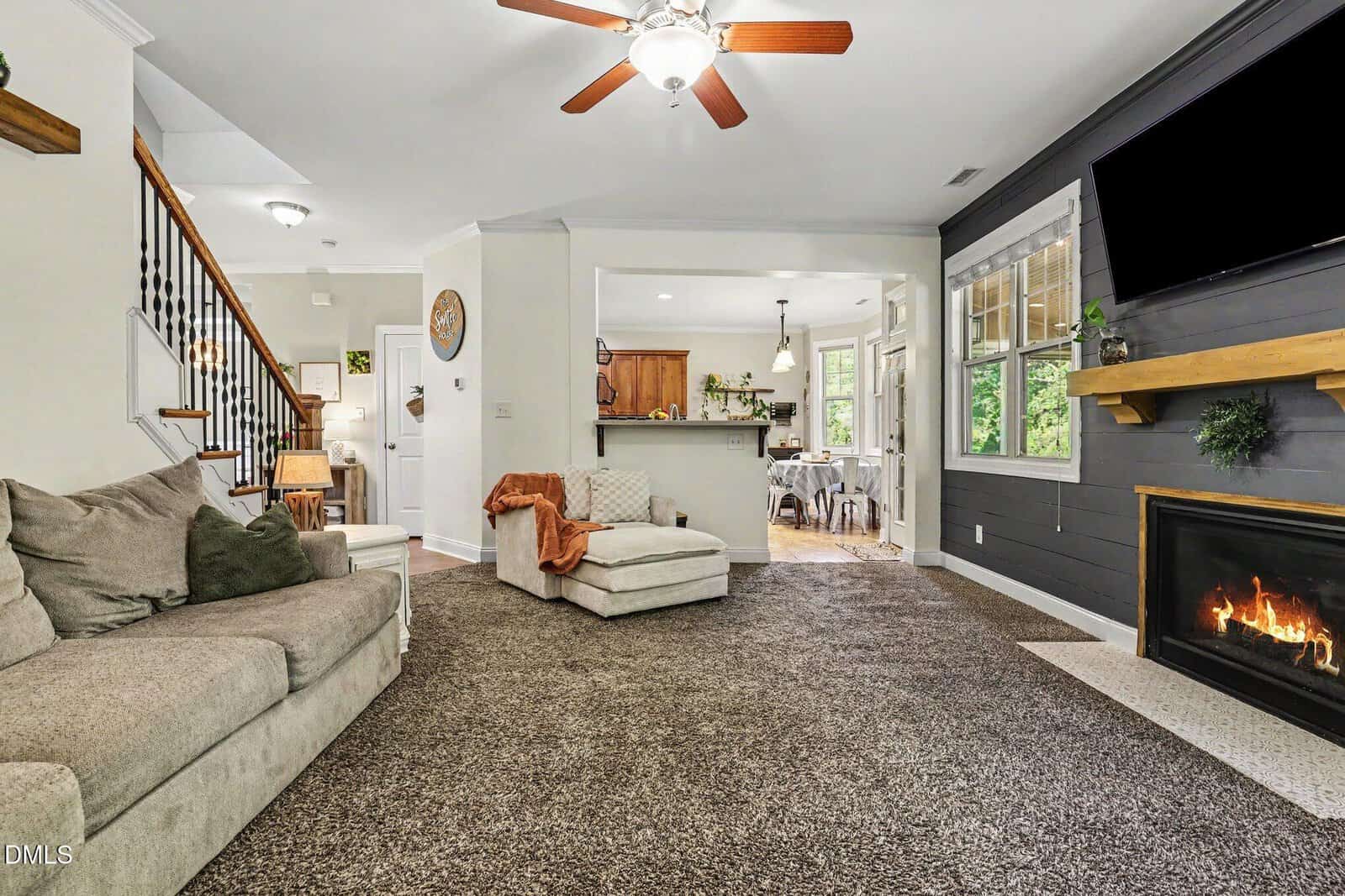 Cozy living room with gray sectional, brown carpet, fireplace, mounted TV, and open view into a bright kitchen. Ceiling fan and staircase are visible, with natural light from large windows.