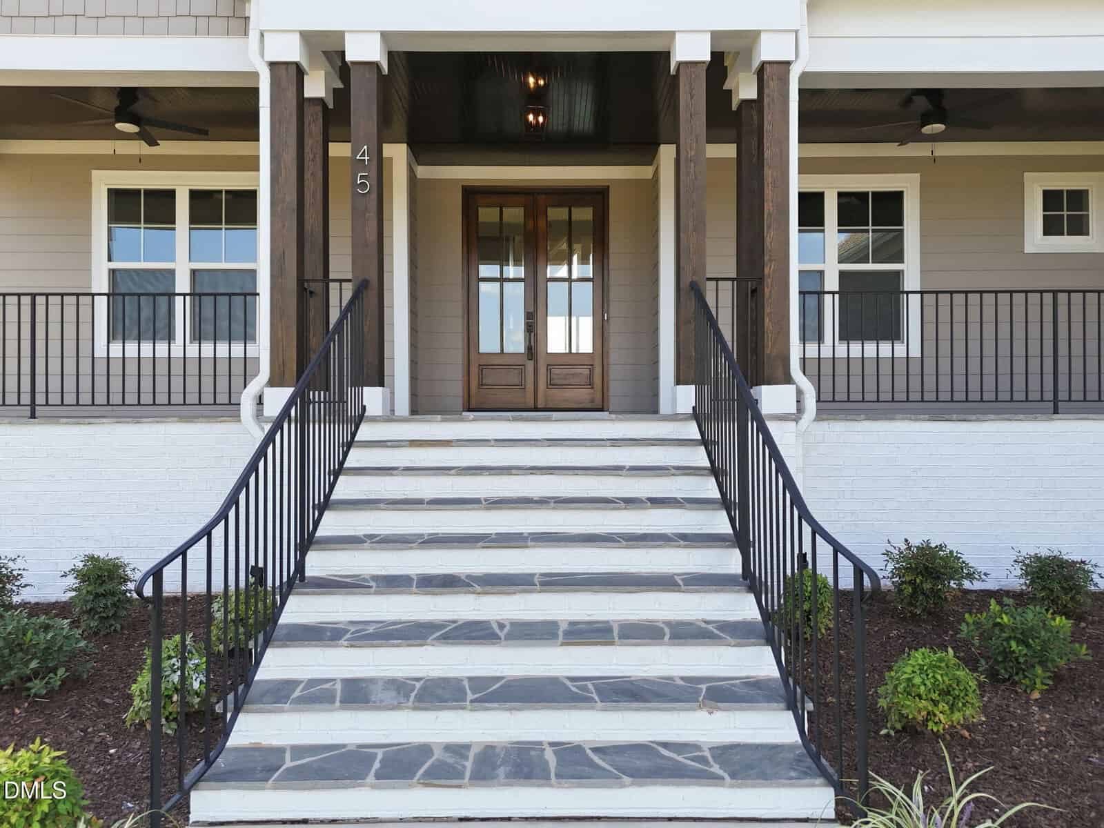 Front steps with black railings lead to double wooden doors of a modern house with tan siding and white trim. Small shrubs are planted along the walkway and steps. The number 45 is visible beside the door.