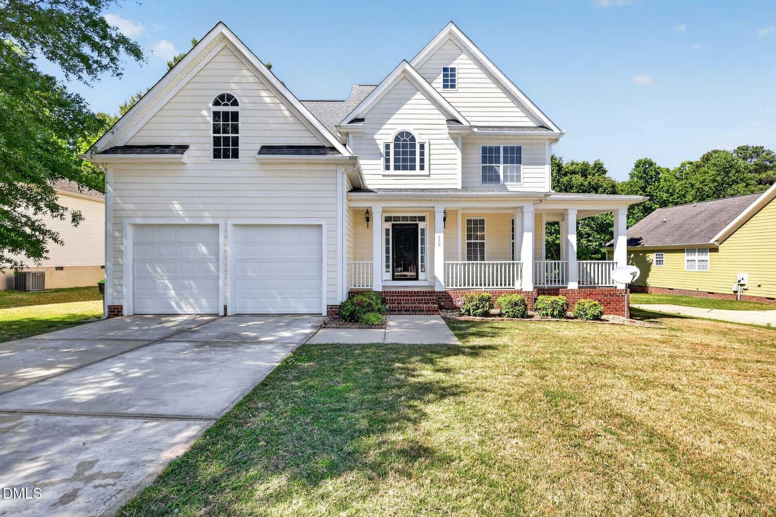 Two-story white house with a covered front porch, two-car garage, and large front lawn. Brick steps lead to the entrance, and green shrubs line the base of the porch. The sky is clear and sunny.