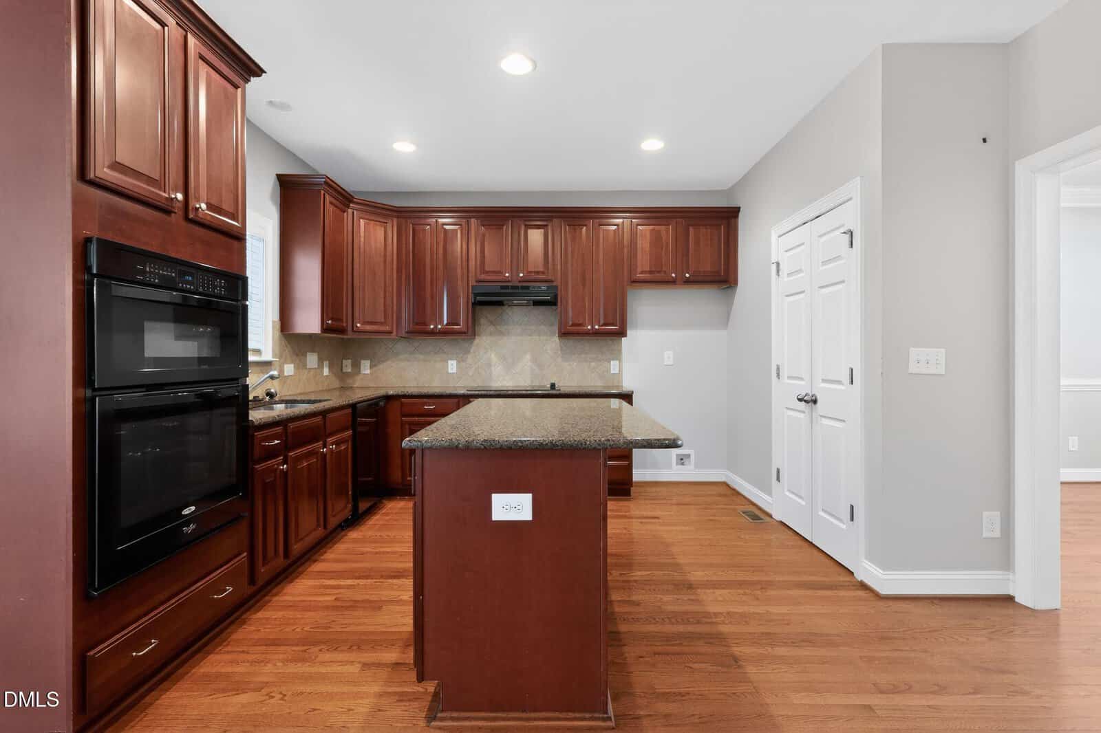 Spacious kitchen with dark wood cabinets, granite countertops, a center island, double wall ovens, and hardwood floors. White paneled double doors and light gray walls add a modern touch. Bright overhead recessed lighting.