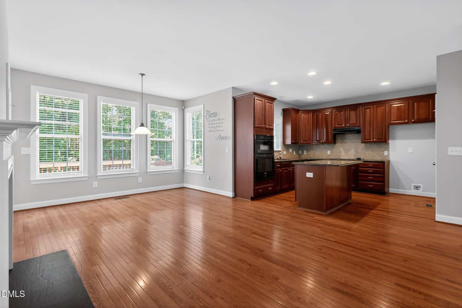 Open-concept kitchen and dining area with hardwood floors, large windows, granite countertops, dark wood cabinets, and an island under recessed lighting. Light gray walls and lots of natural light fill the space.
