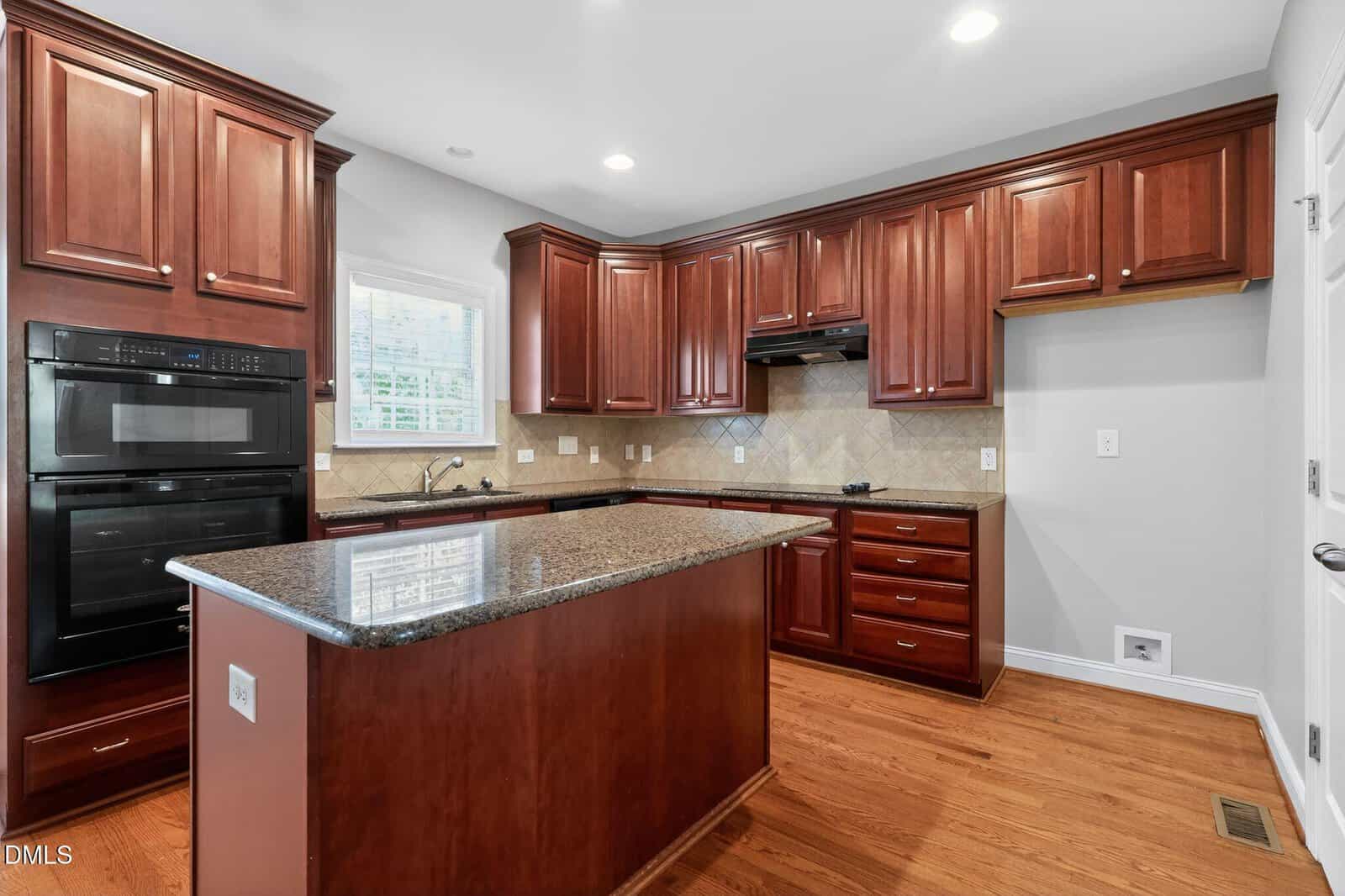 Modern kitchen with dark wood cabinets, granite countertops, island, double oven, stove, tile backsplash, under-cabinet lighting, hardwood floors, and a window above the sink.