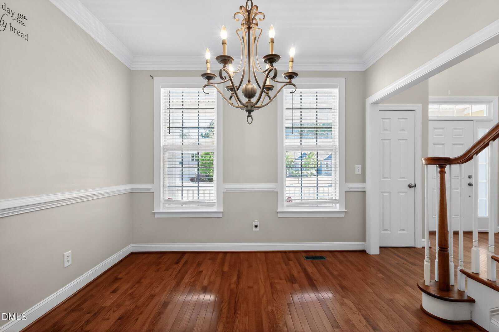 A dining room with light beige walls, white trim, two large windows with blinds, a chandelier, and polished hardwood floors. A staircase with a wooden handrail is visible on the right.